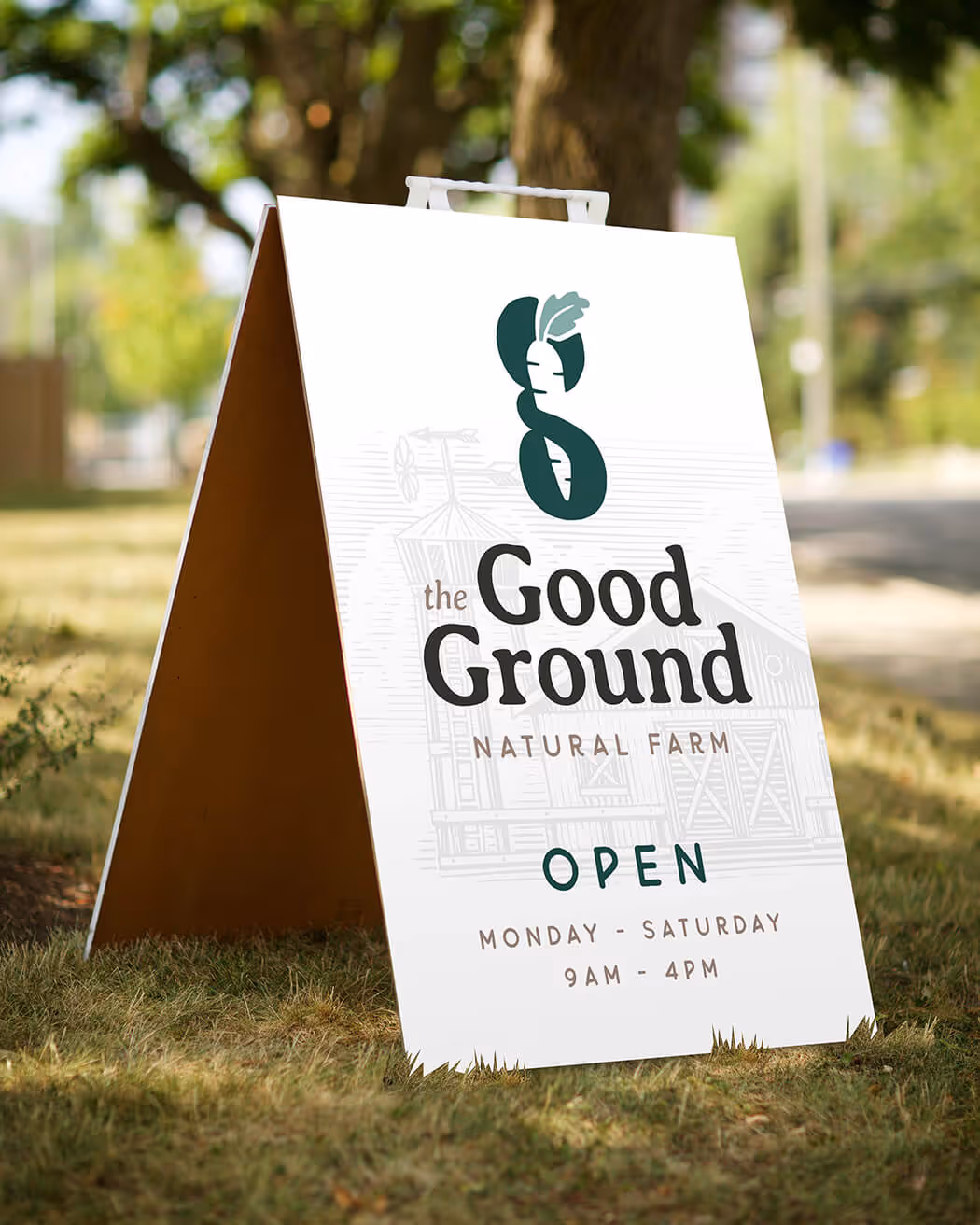 A-frame sign on grass displaying 'The Good Ground Natural Farm' logo with a carrot illustration and hours open Monday to Saturday 9AM to 4PM.
