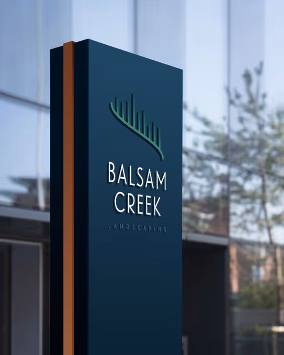 Tall, dark blue sign with a green leaf logo and white text reading 'Balsam Creek Landscaping' in front of a glass building.