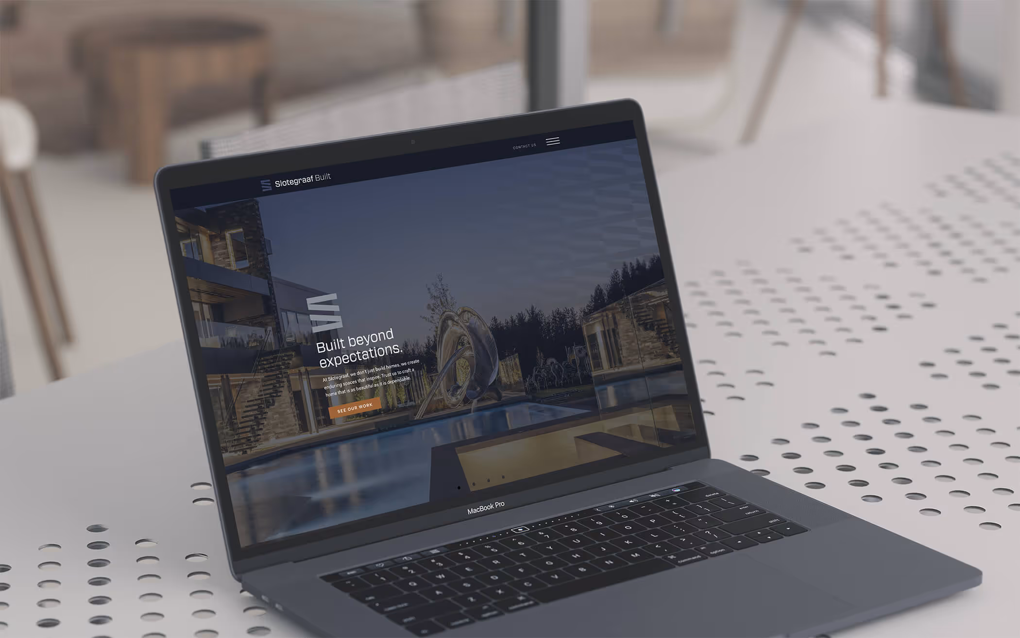 MacBook Pro laptop on a perforated white table displaying the Slotegraaf Built website homepage with a modern house and pool background.