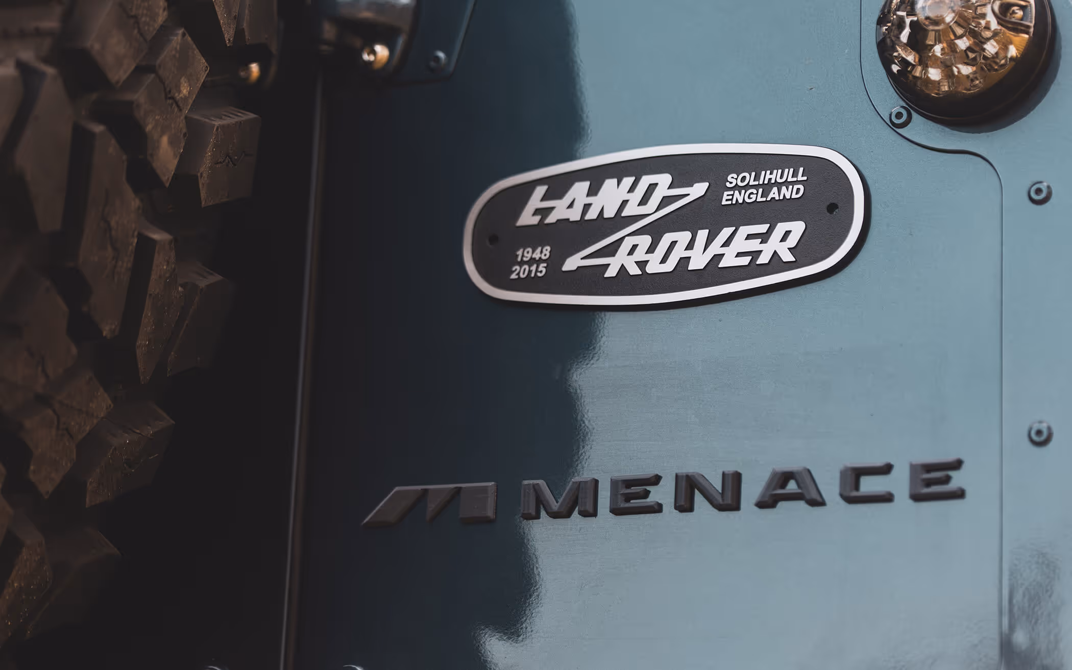 Close-up of a Land Rover vehicle badge and the word 'MENACE' on a blue-green body panel next to a rugged tire.