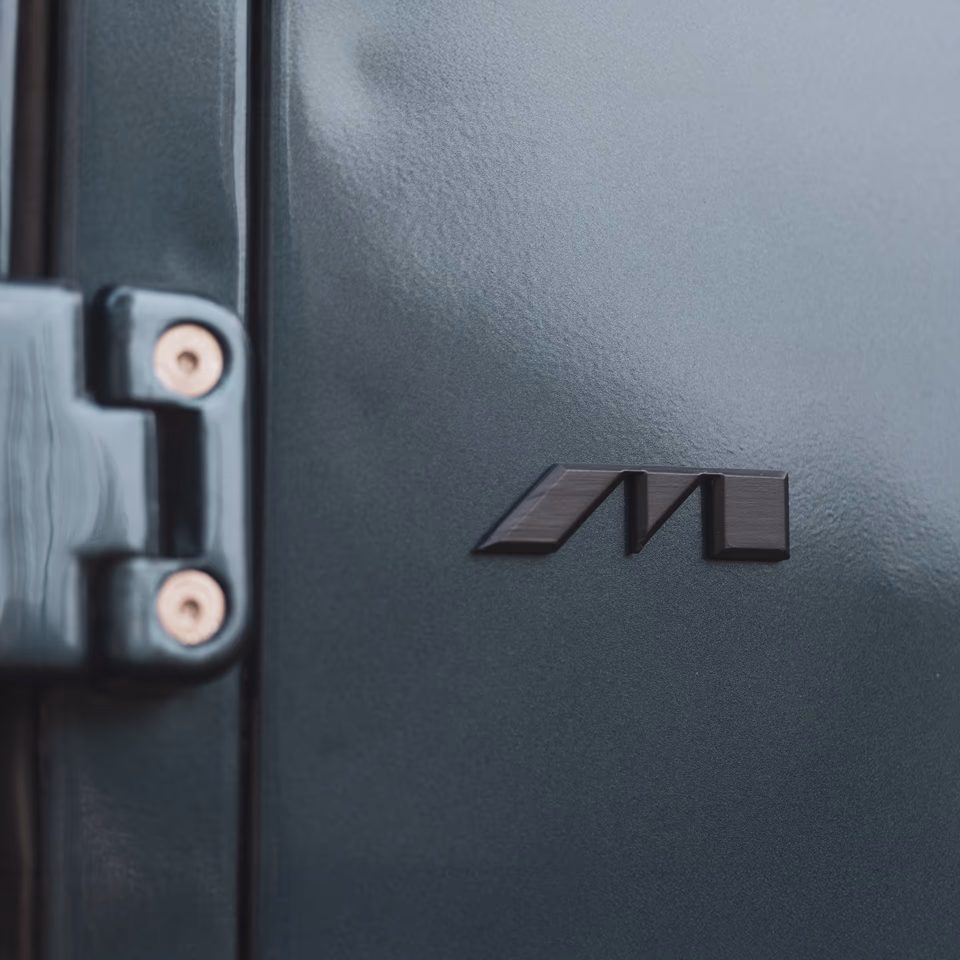 Close-up of a dark green metal surface with a black emblem resembling the letters 'MT' and part of a hinge with screws.