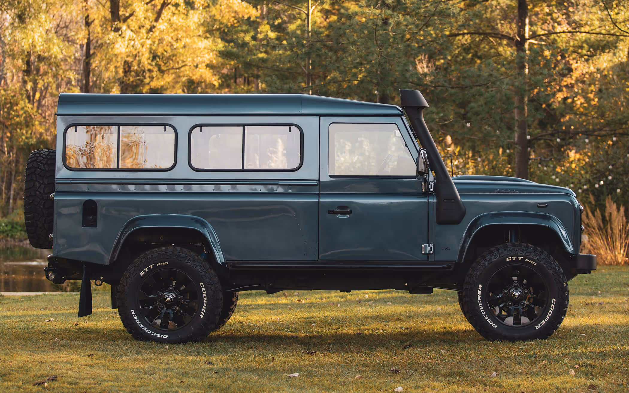 Side view of a dark green Land Rover Defender parked on grass with trees and a pond in the background.