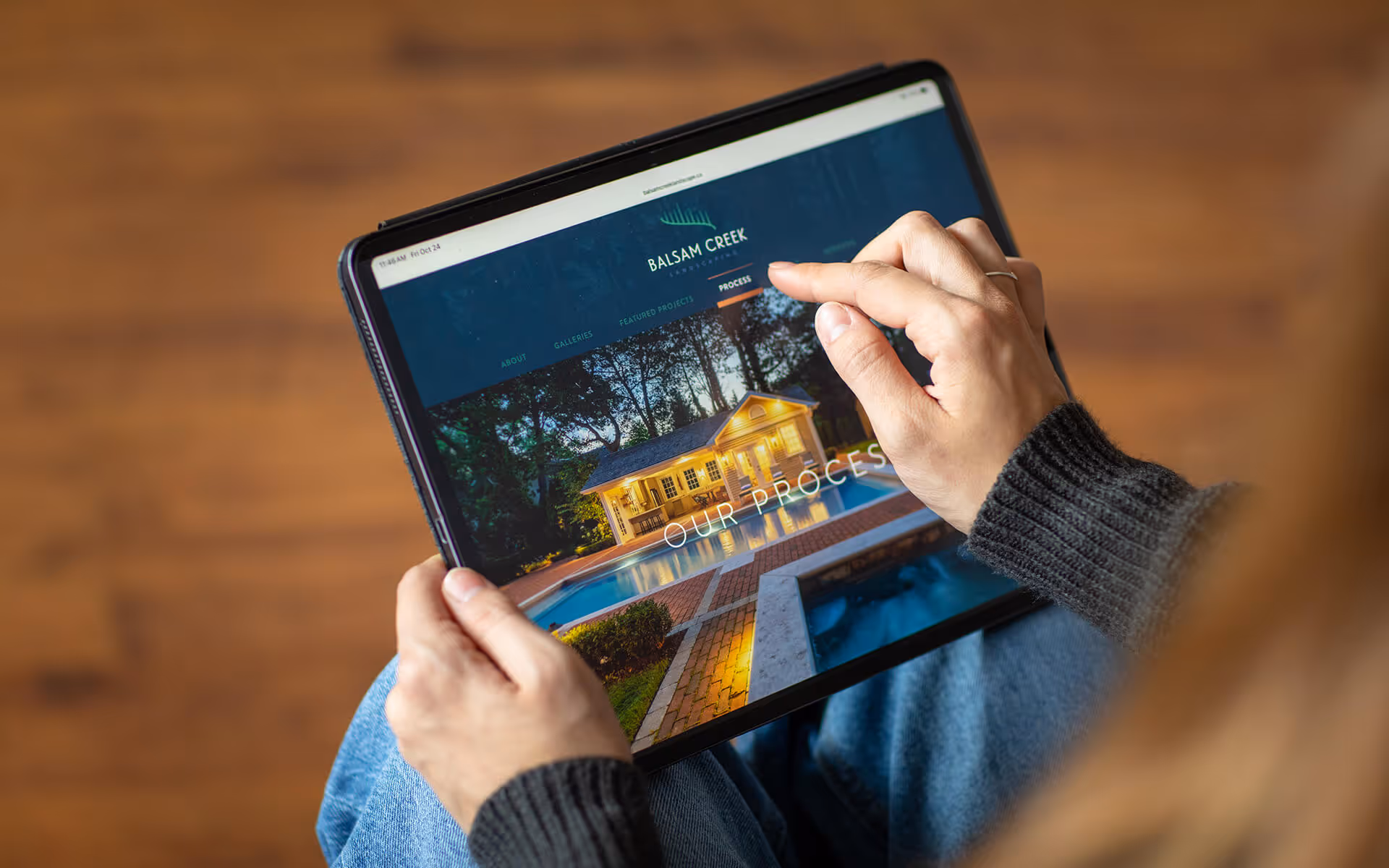 Person holding a tablet displaying the Balsam Creek Landscape website showing a nighttime backyard with a lit house and pool under the 'Our Process' section.