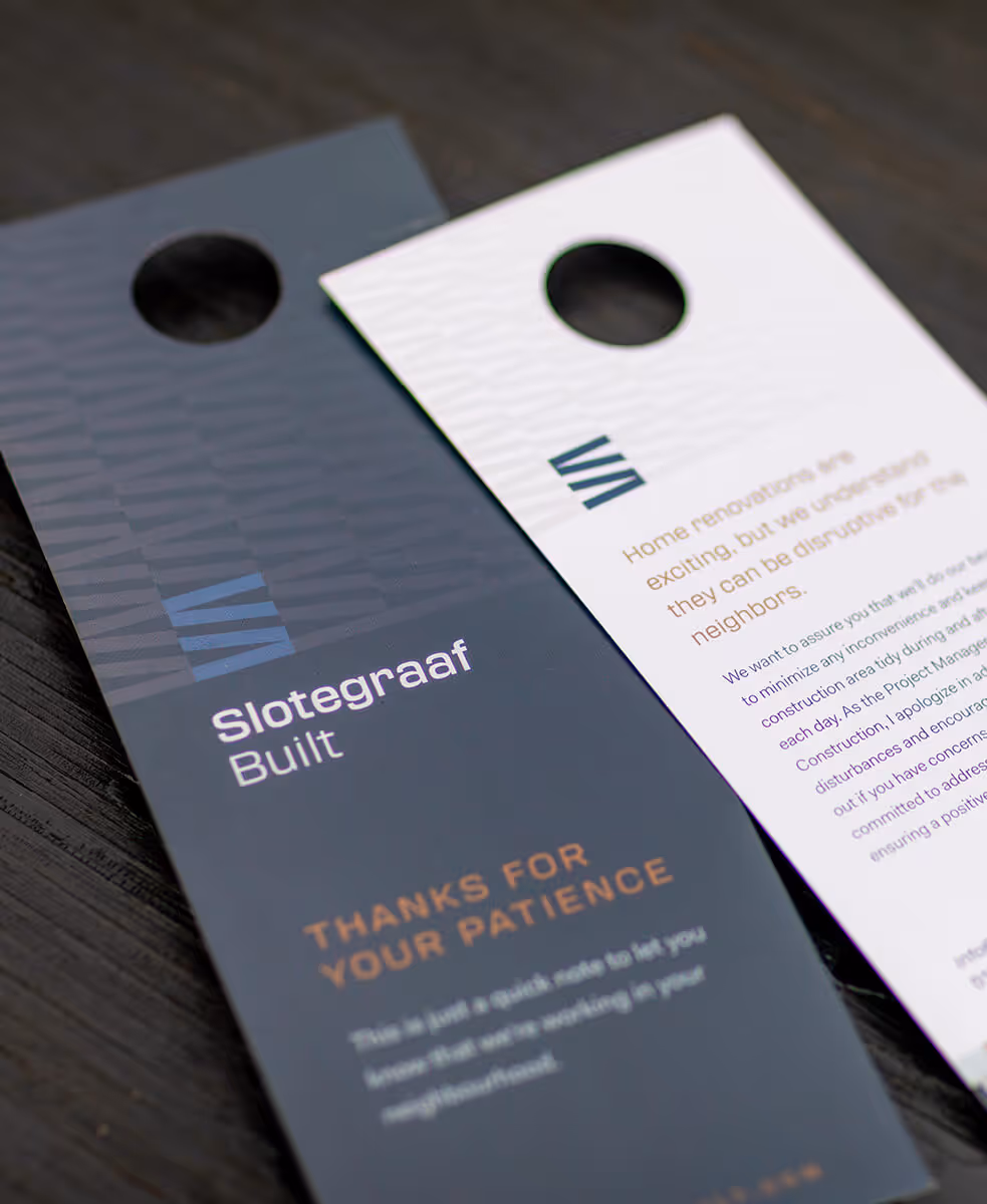 Two door hangers on a dark wooden surface, one black with the text 'Slotegraaf Built' and 'Thanks for Your Patience,' the other white with a message about home renovations.