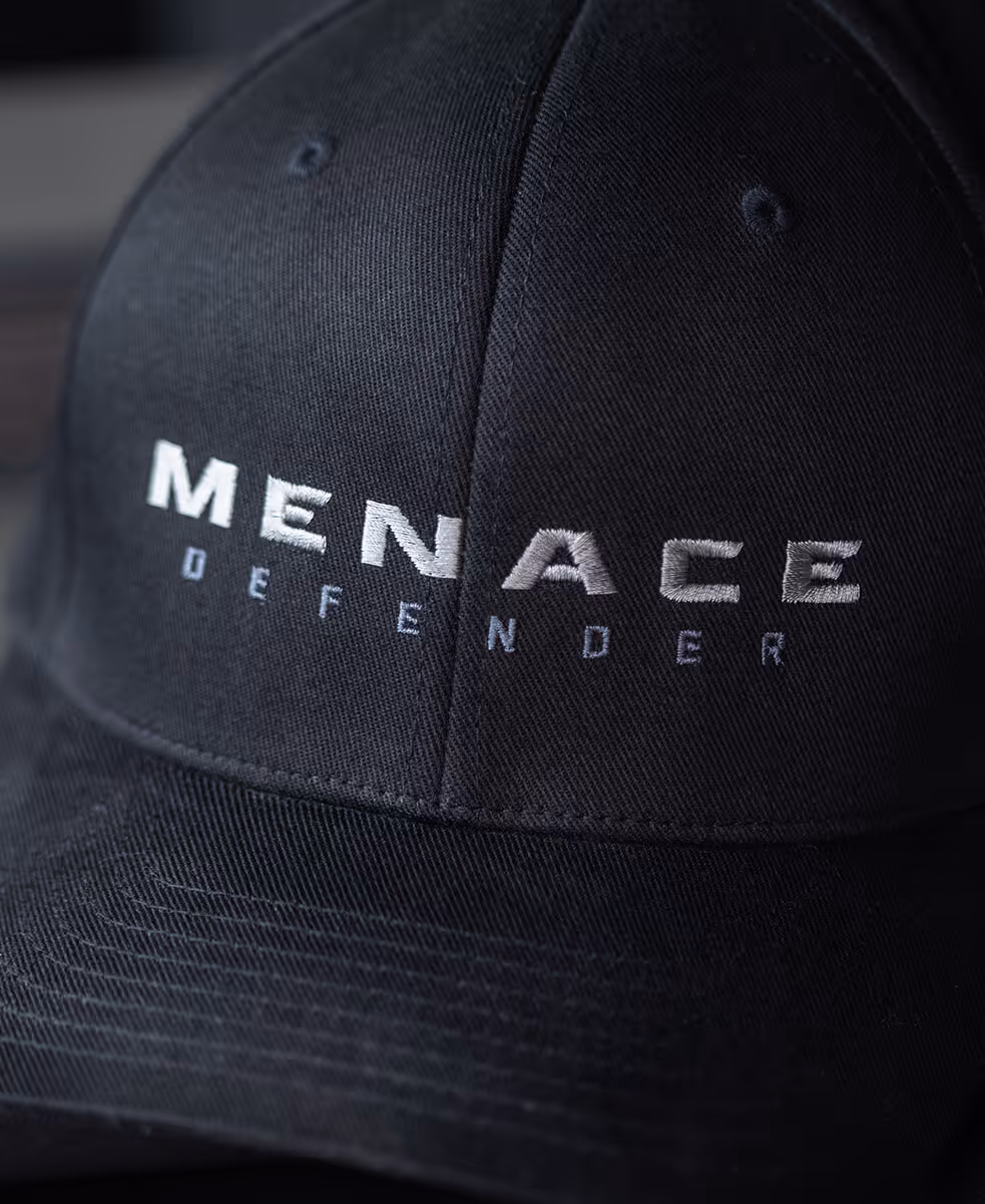 Close-up of black cap with white embroidered text saying 'MENACE DEFENDER'.