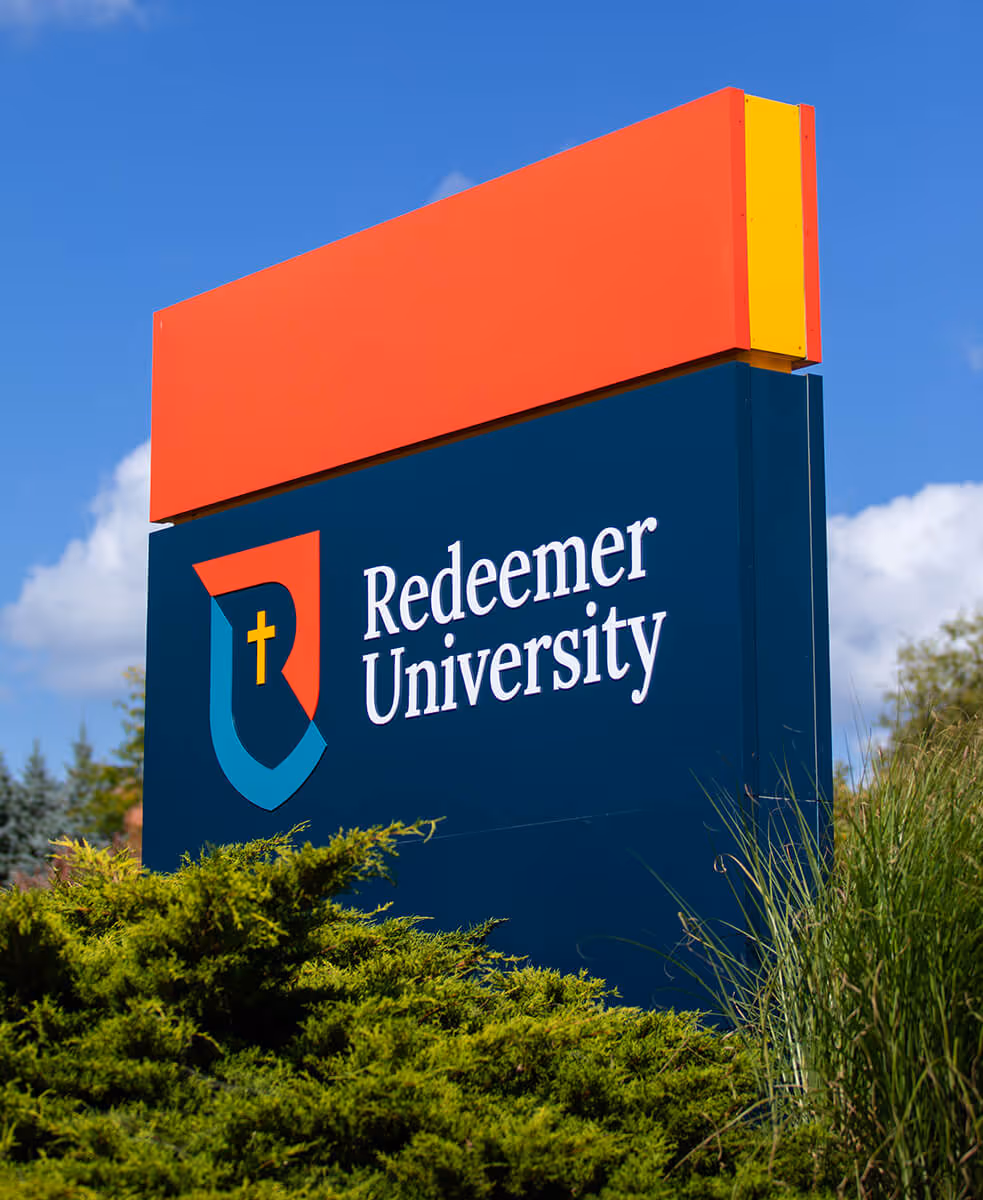 Outdoor sign for Redeemer University with a blue background, red top section, and university logo featuring a cross.