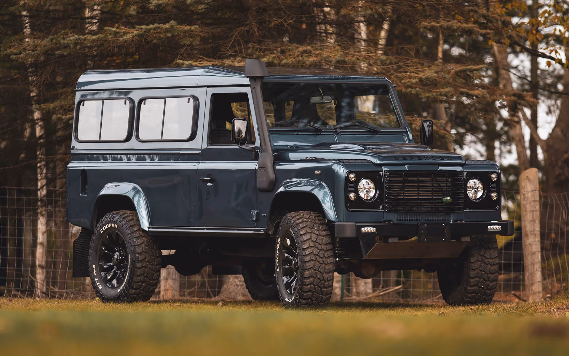 Dark blue Land Rover Defender parked on grass with forest background.