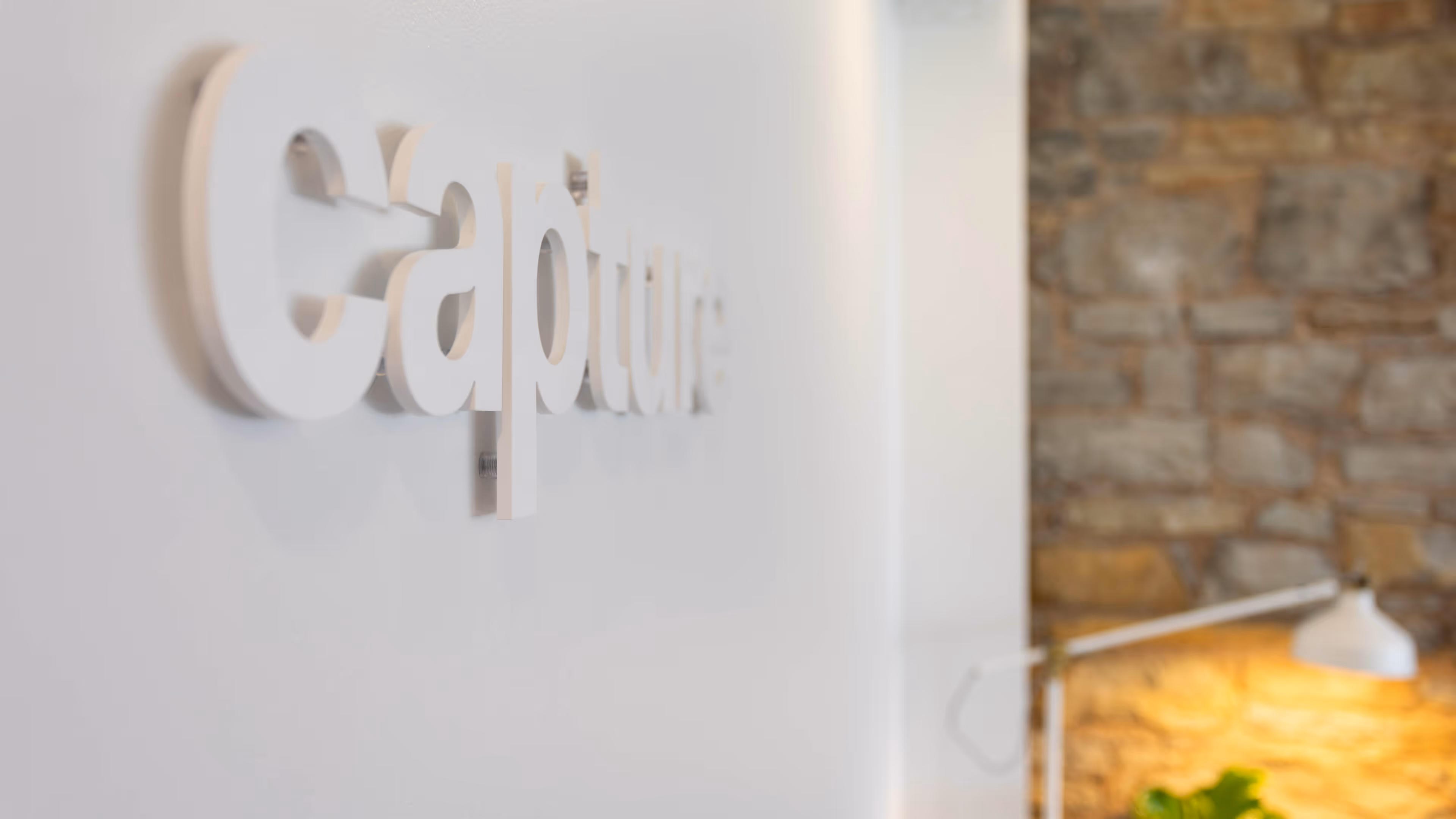White 3D letters spelling 'Capture' mounted on a white wall with a stone wall and a white desk lamp in the background.