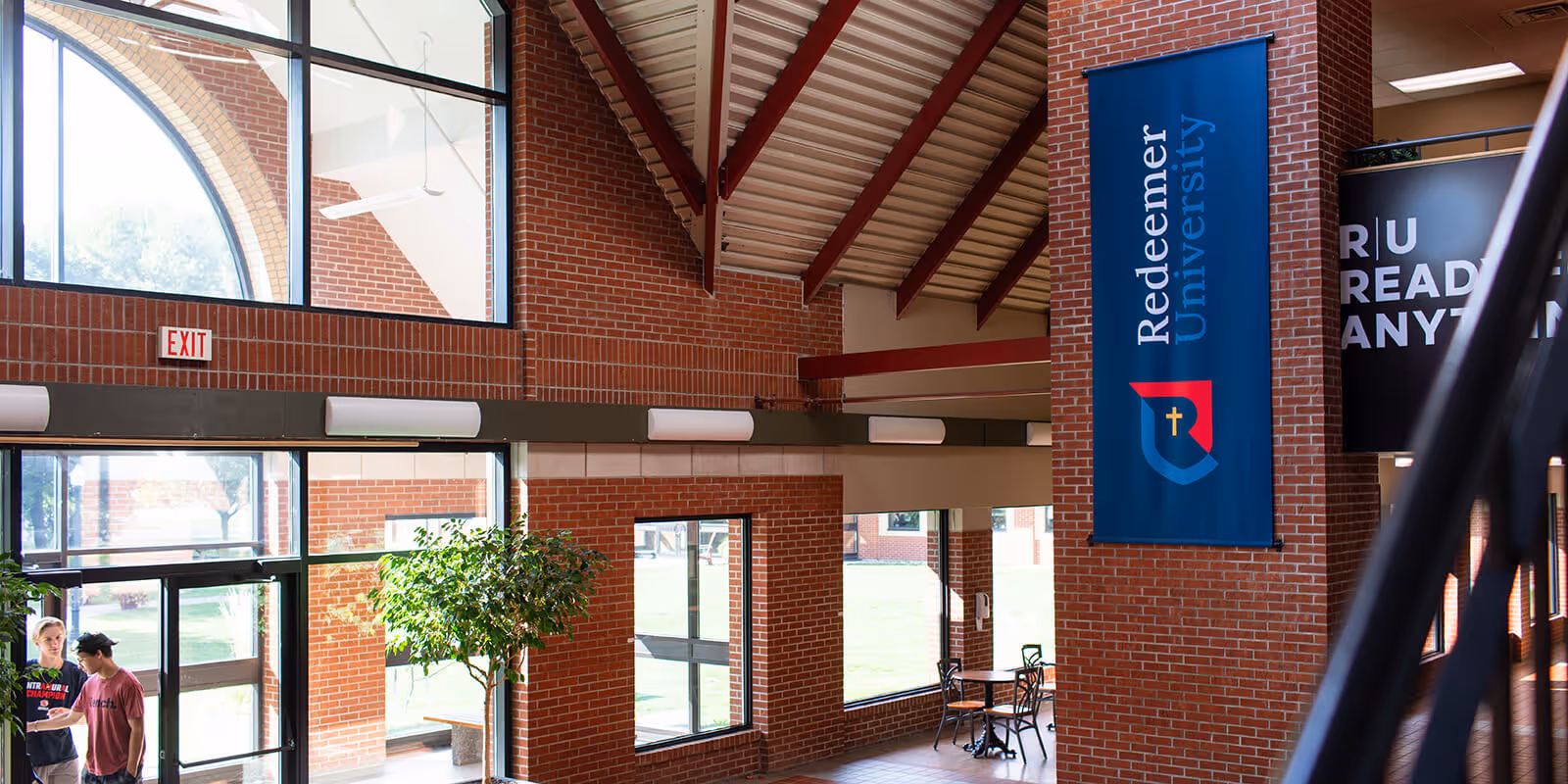Interior of a university building with large windows, a Redeemer University banner on a brick pillar, and two students standing near the entrance.