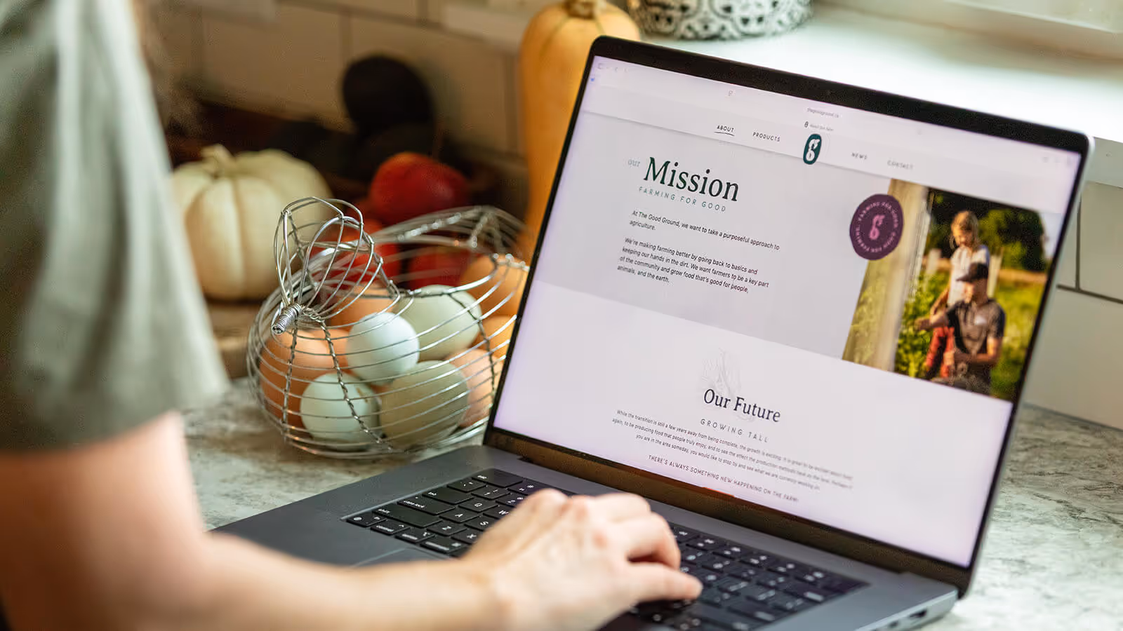 Person using a laptop displaying a website with 'Mission' text about farming, next to a wire basket of eggs on a kitchen counter.