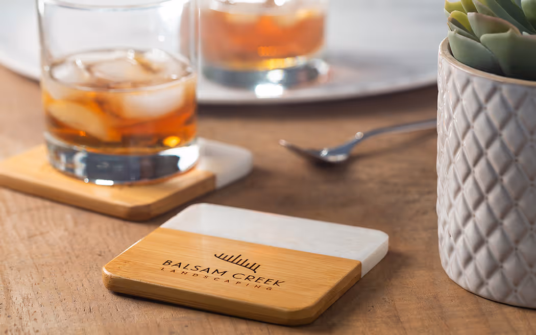 Rectangular coaster made of wood and white stone with Balsam Creek Landscaping logo on a wooden table next to a plant pot and a glass of iced drink.