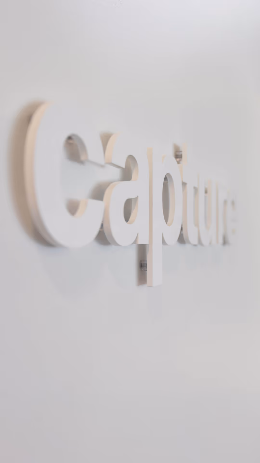 Close-up of white 3D letters spelling “Capture” mounted on a light gray wall.