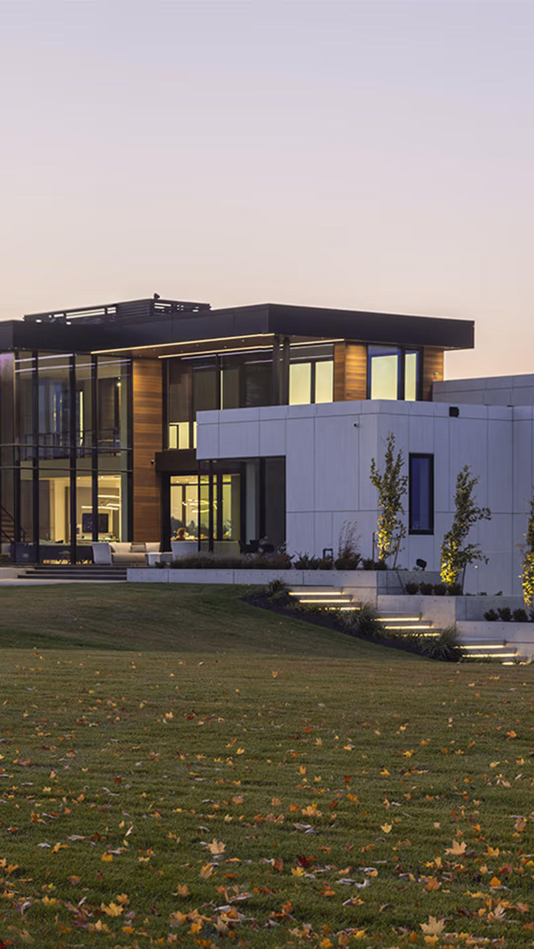 Modern two-story house with large glass windows, illuminated stairs, and a lawn scattered with autumn leaves at dusk.