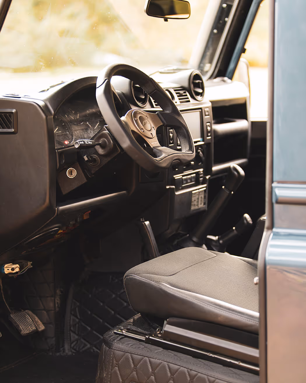 Interior of a vehicle showing a textured steering wheel, dashboard controls, and a fabric driver seat with quilted floor mats.