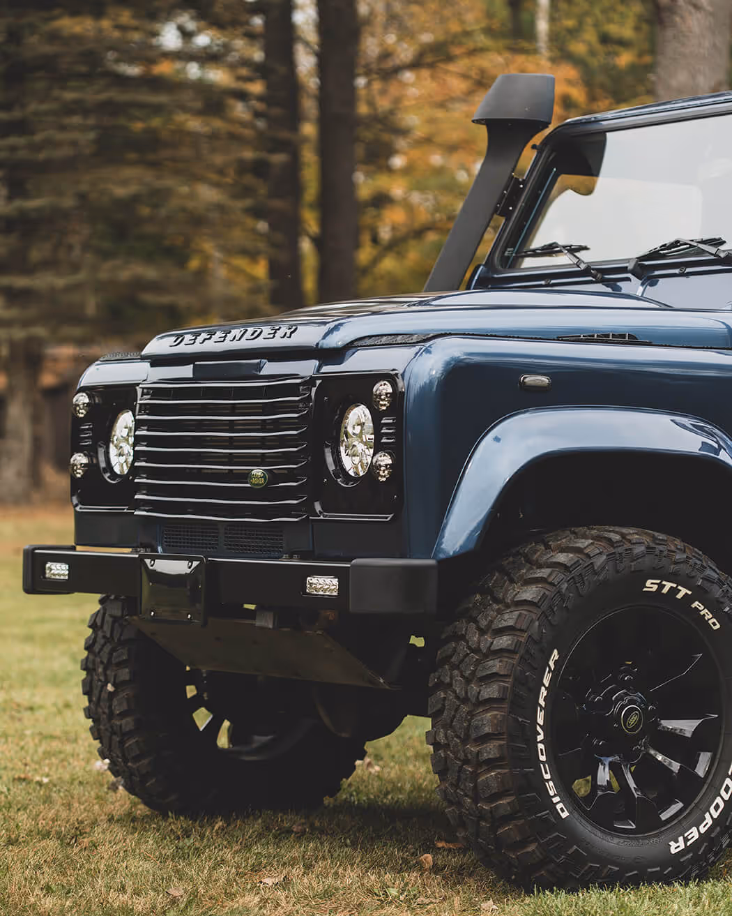 Front side view of a blue Land Rover Defender with rugged off-road tires parked on grass.