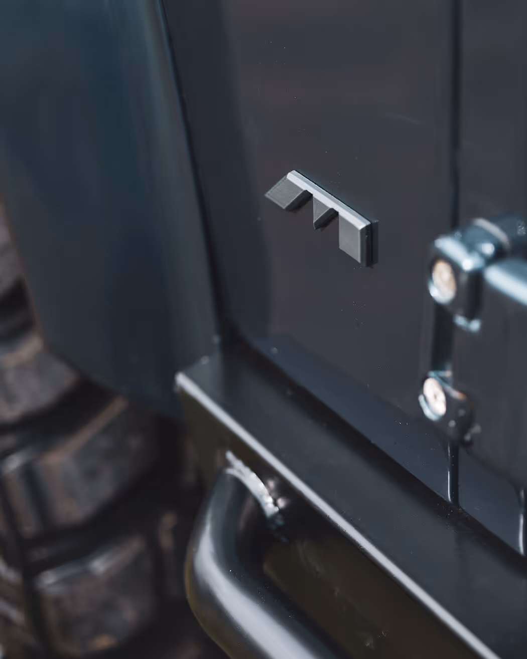 Close-up of a black vehicle's rear corner showing a metal handle, hinge, and a small emblem with three geometric shapes.