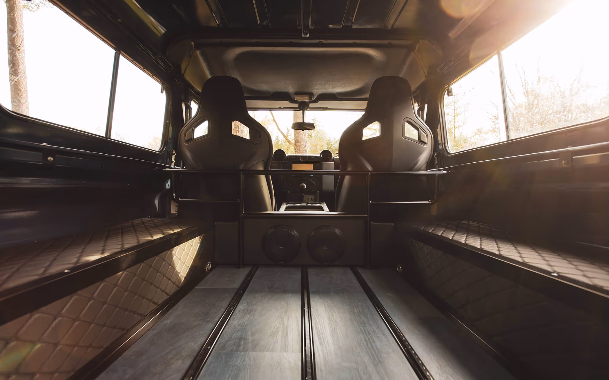 Interior of a vehicle showing two black bucket seats, quilted side benches, and a wooden floor with metal tracks.