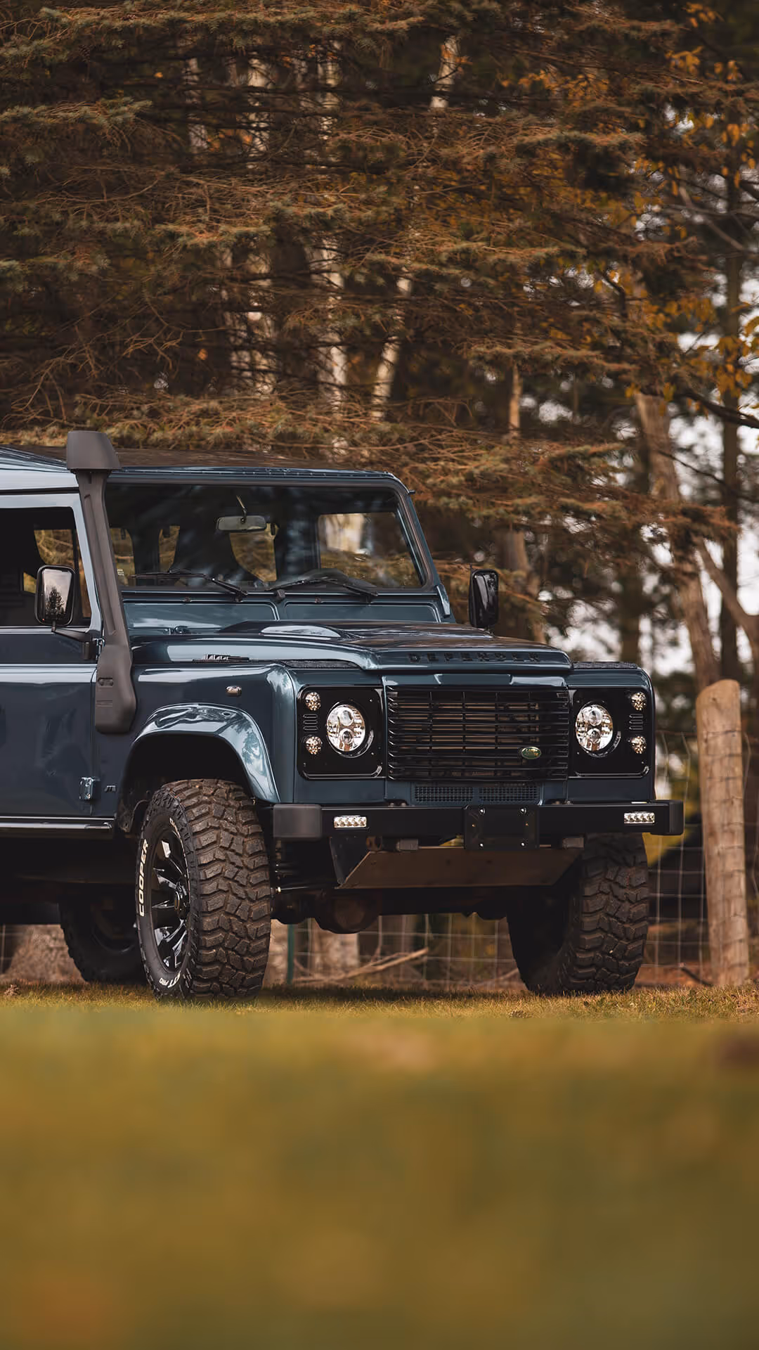 Front side view of a dark green Land Rover Defender parked on grass with trees in the background.