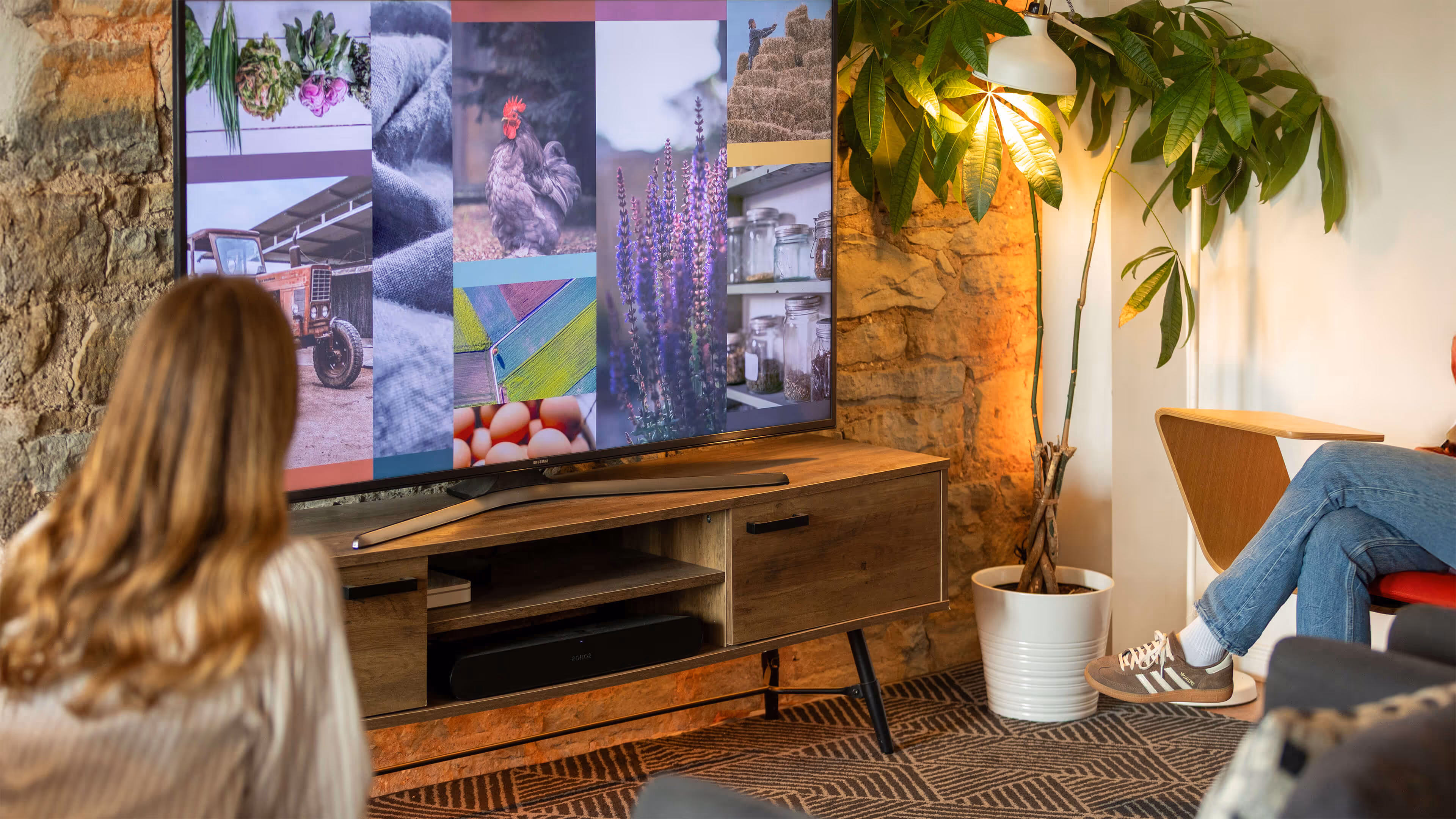 Two people sitting in a cozy living room watching a large TV screen displaying images of farm produce, animals, and nature.