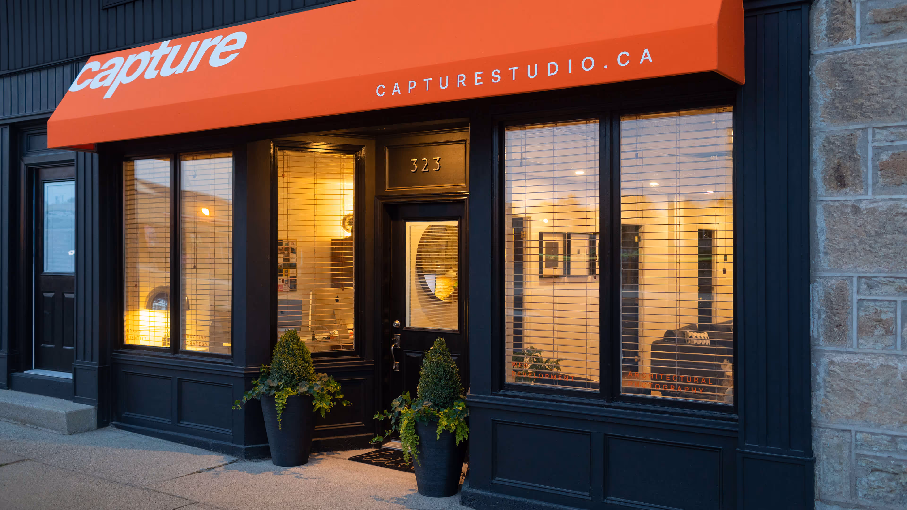 Storefront of Capture Studio with black framed windows and door, orange awning, and two large potted plants at the entrance.