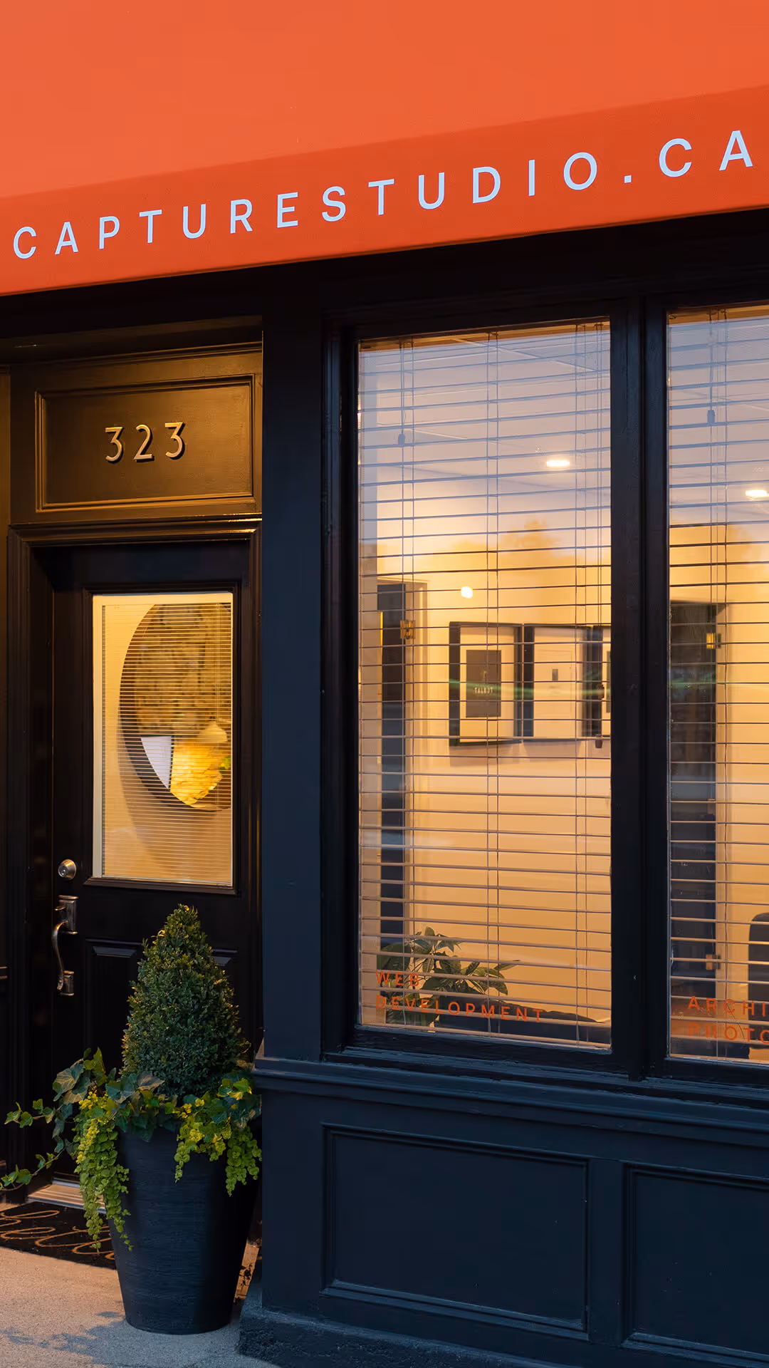 Entrance of a black-painted office with the number 323, a potted plant, and windows with blinds showing framed pictures inside.
