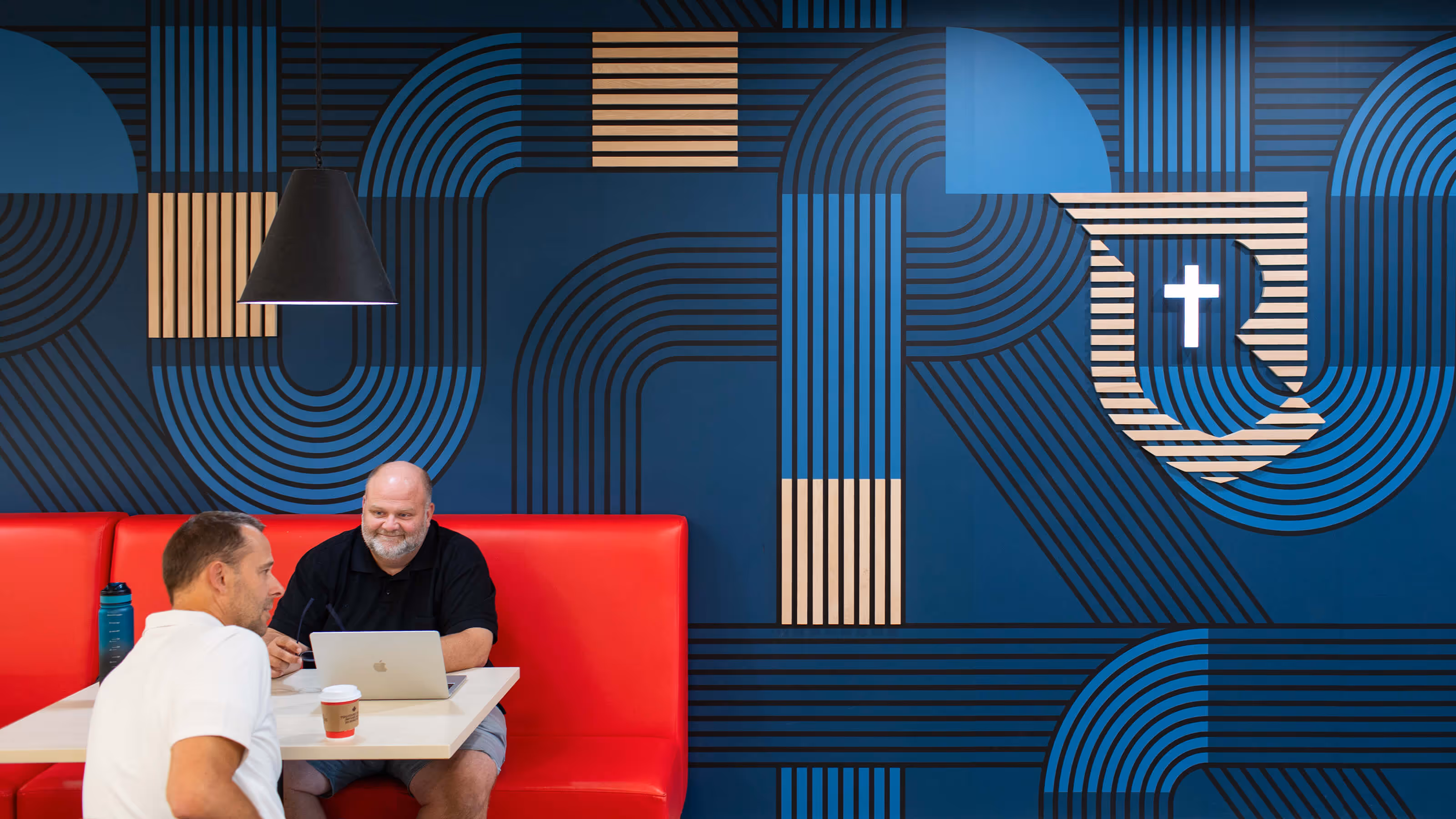 Two men sitting at a white table with red booth seating, one using a laptop, against a blue wall with modern black and beige geometric patterns and a shield logo with a white cross.