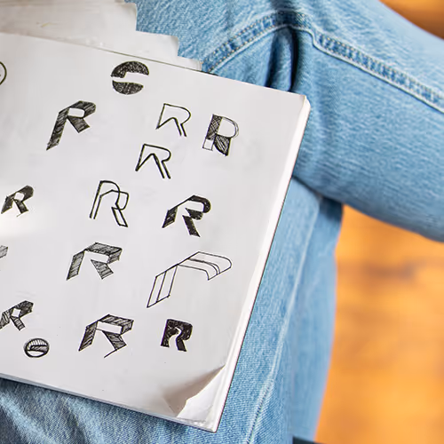 Sketchbook page resting on crossed denim-clad legs, displaying various stylized 'R' letter designs and shapes.