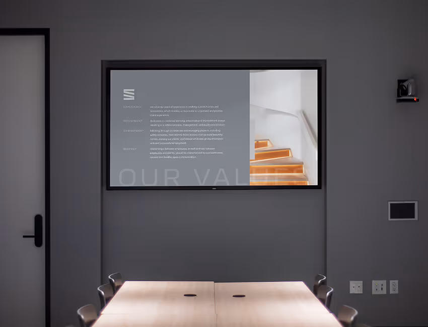 Conference room with table and chairs facing a wall-mounted screen displaying text about values alongside a photo of wooden stairs.