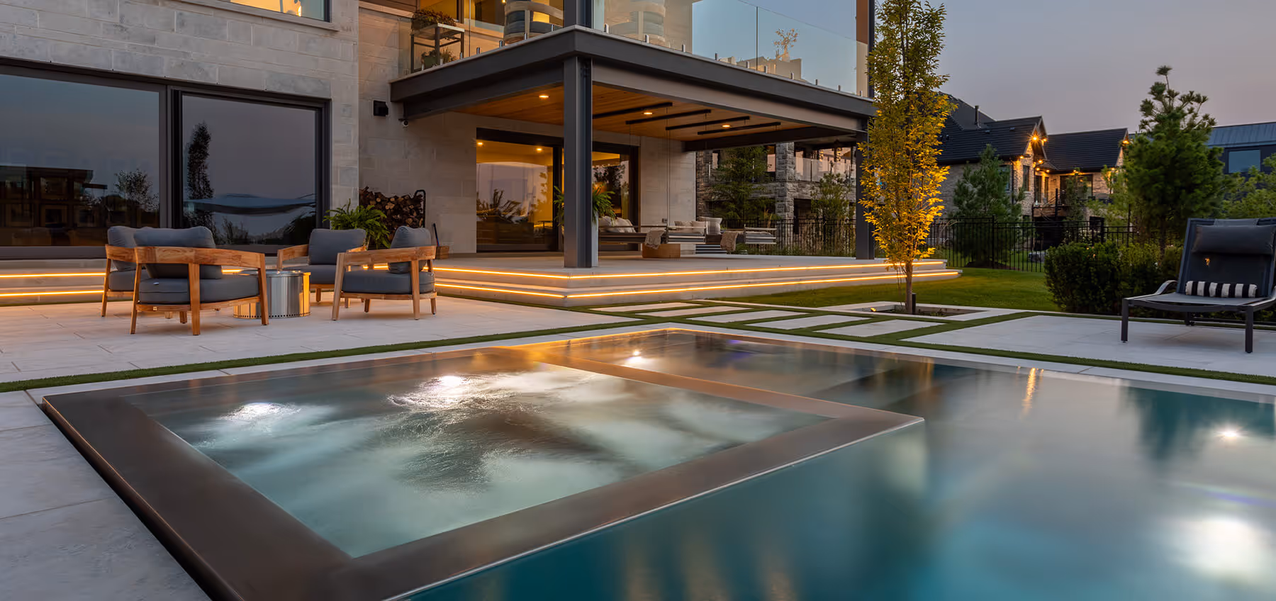 Modern backyard patio with jacuzzi, swimming pool, patio seating, and illuminated steps at dusk.
