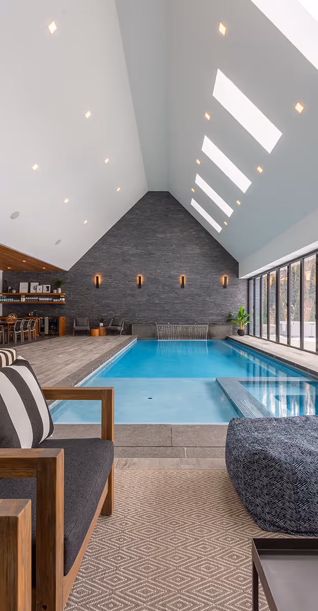 Indoor swimming pool with light blue water, modern seating, large windows, and high ceiling with recessed lights and skylights.
