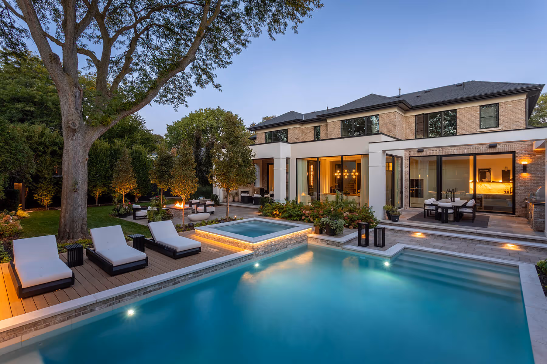 Modern two-story house with a lit swimming pool, hot tub, lounge chairs, and a garden with outdoor seating and fire pit.