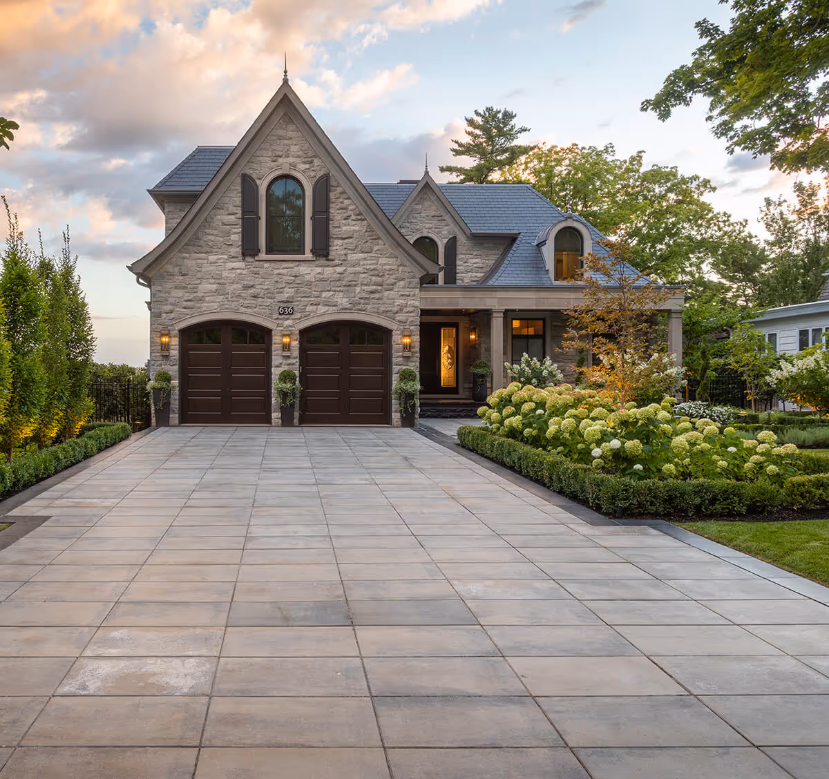 Stone house with two dark brown garage doors, a large paved driveway, and landscaped garden with green shrubs and flowers.