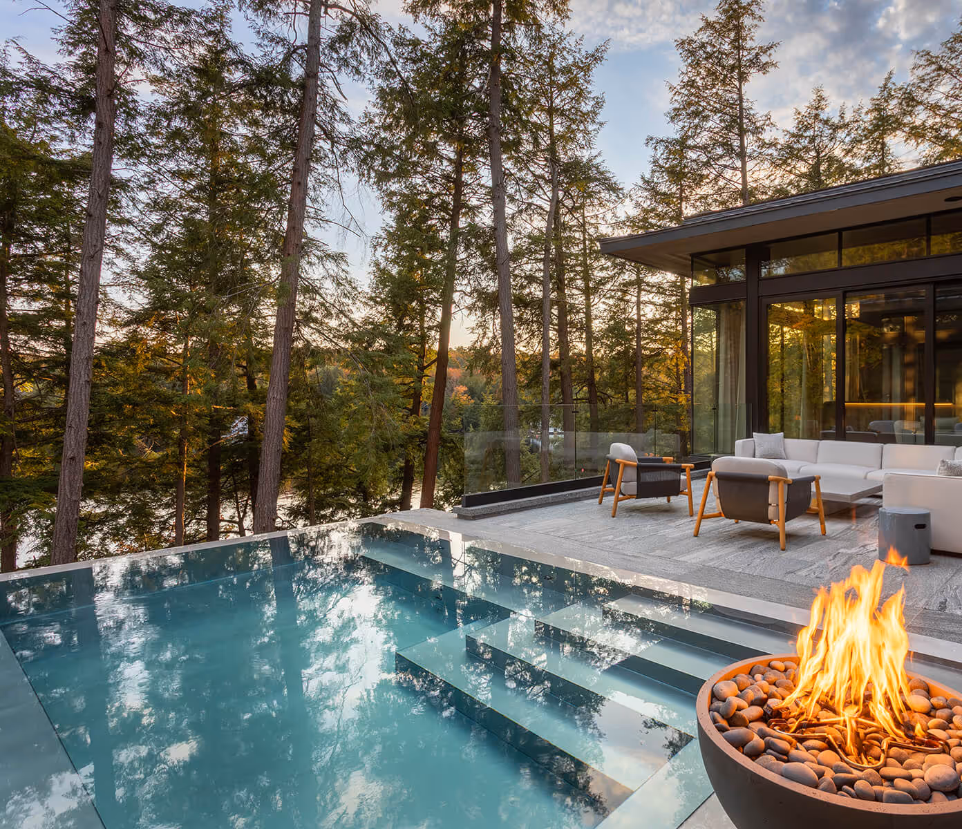 Modern outdoor patio with sectional sofa, chairs, infinity pool, and fire pit surrounded by tall pine trees.