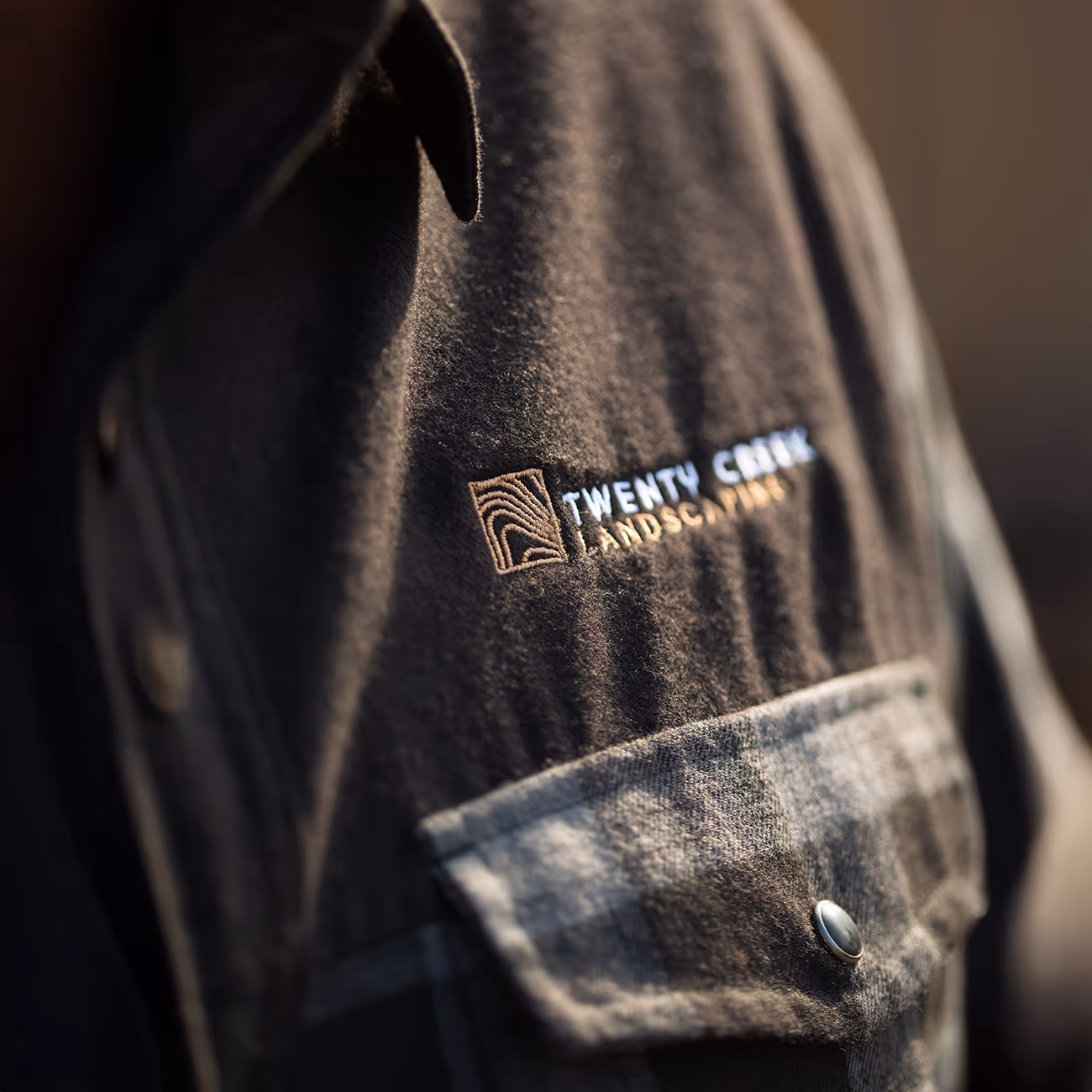 Close-up of a black jacket with a checked pocket and embroidered logo reading 'Twenty Creek Landscaping'.