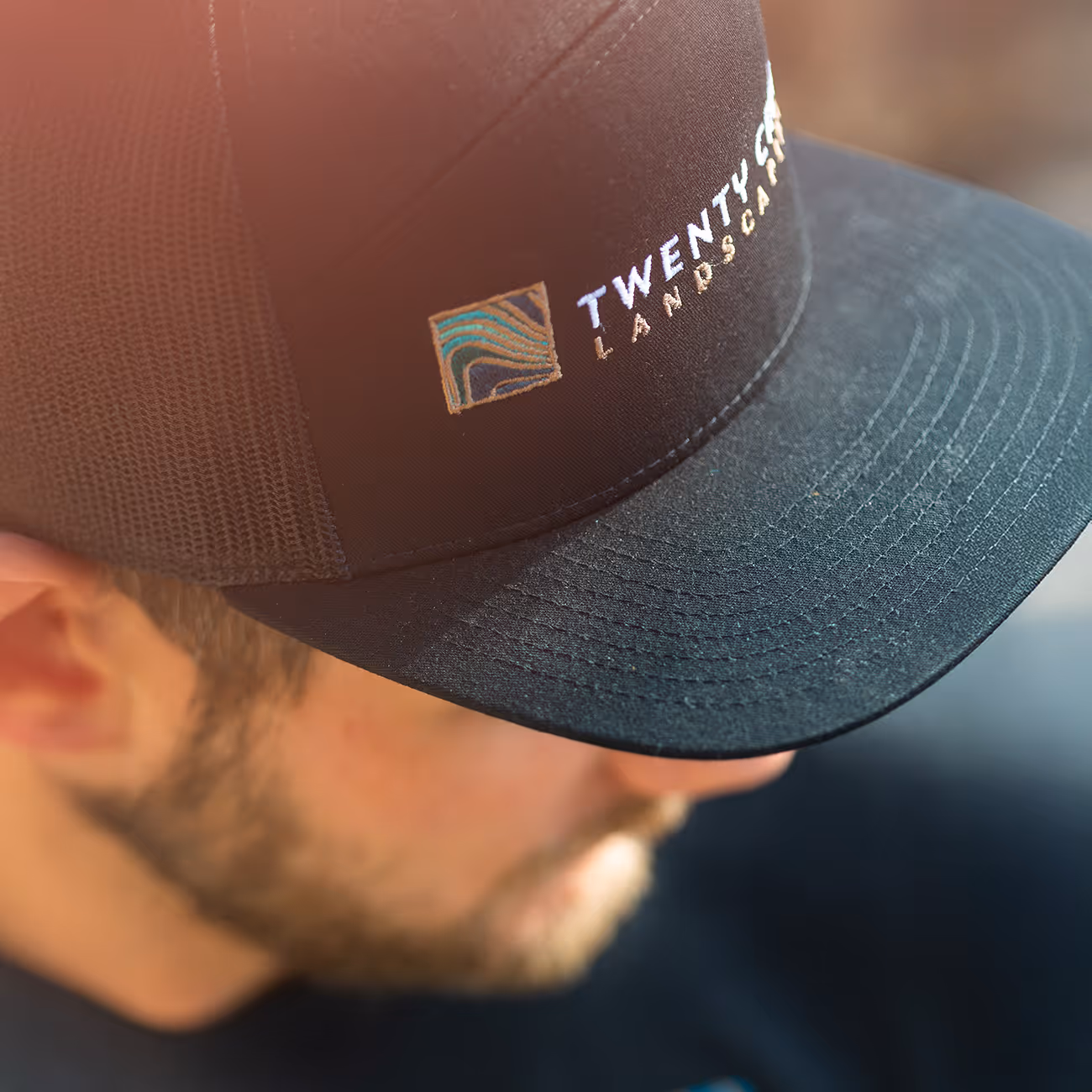 Close-up of a man wearing a black mesh trucker hat embroidered with a multicolored square logo and the text 'TWENTY SIX LANDSCAPING'.
