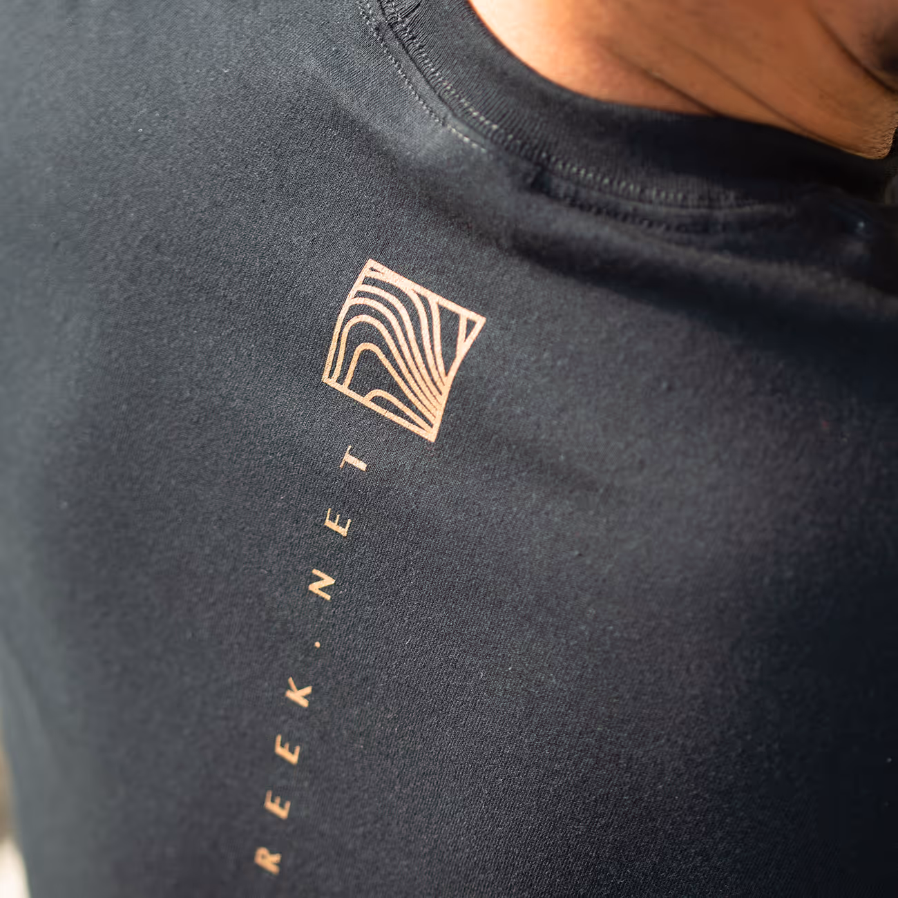 Close-up of a black T-shirt with a gold wavy square logo and the text 'REEK.NET' printed vertically below it.
