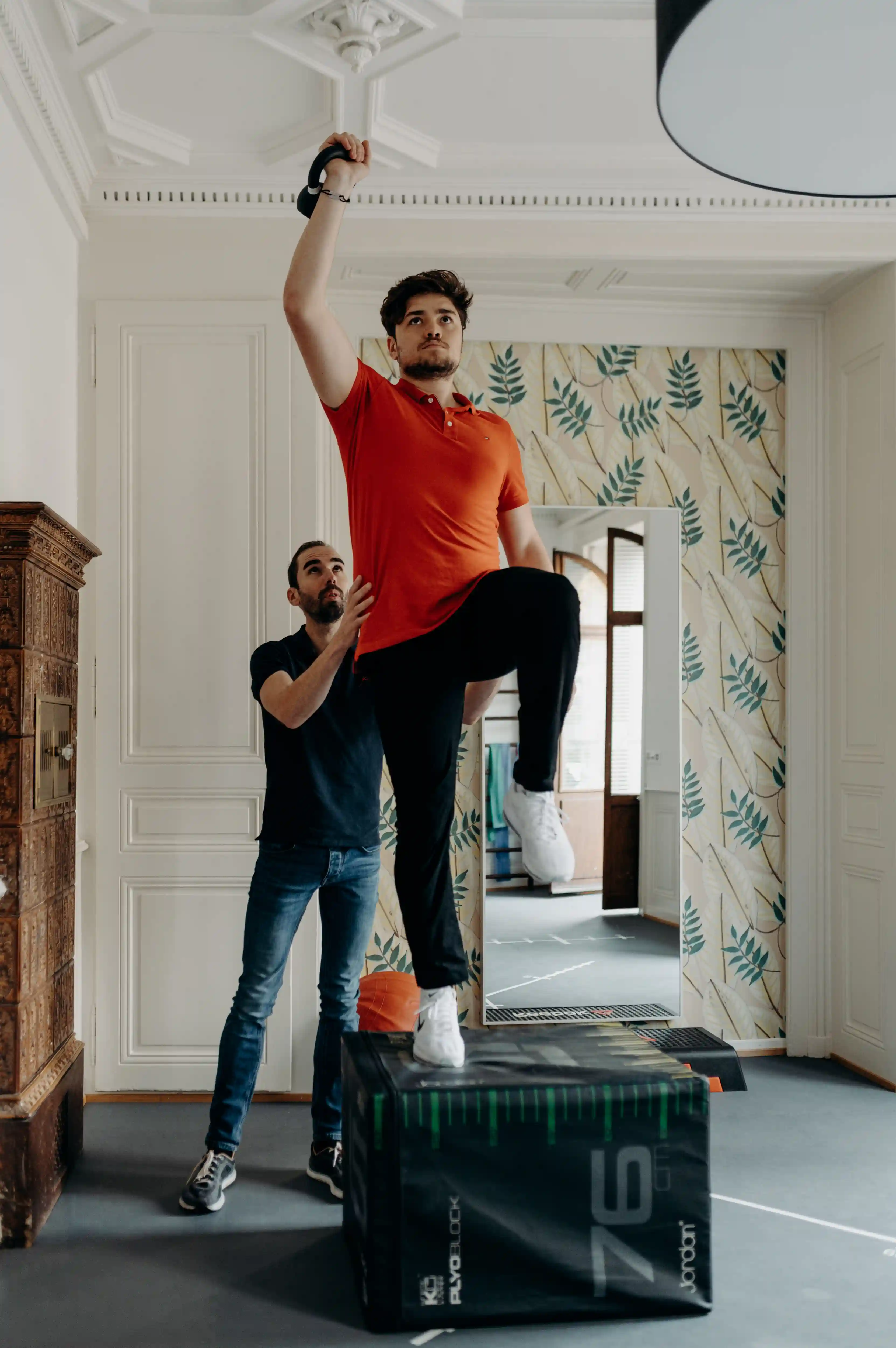 A man in an orange shirt performing a step-up exercise on a large plyometric box while holding a kettlebell overhead, assisted by a trainer in a black shirt.