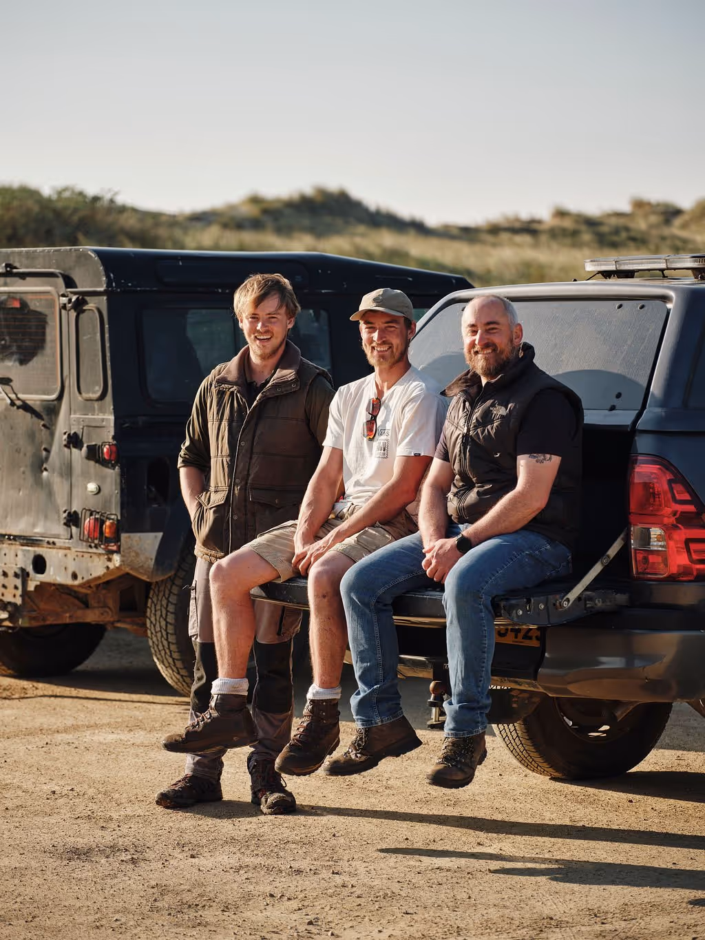 Three men smiling and sitting on the open tailgate of a black off-road vehicle on a dirt road.