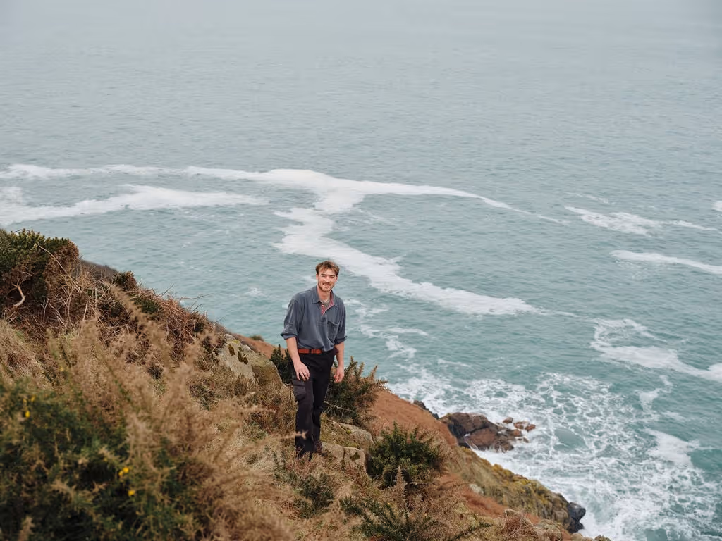 Man smiling while standing on a grassy cliff overlooking the ocean with waves crashing below.
