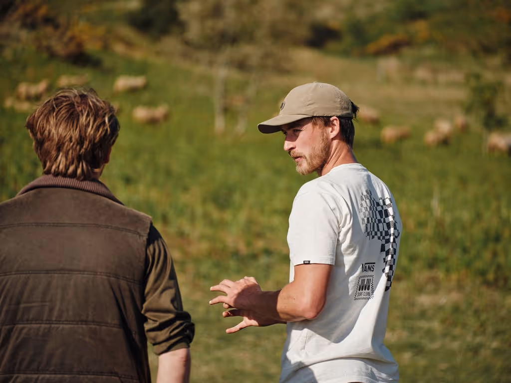 Two men outdoors in a green field, one facing away and the other wearing a cap and white t-shirt with a design, gesturing with his hands.