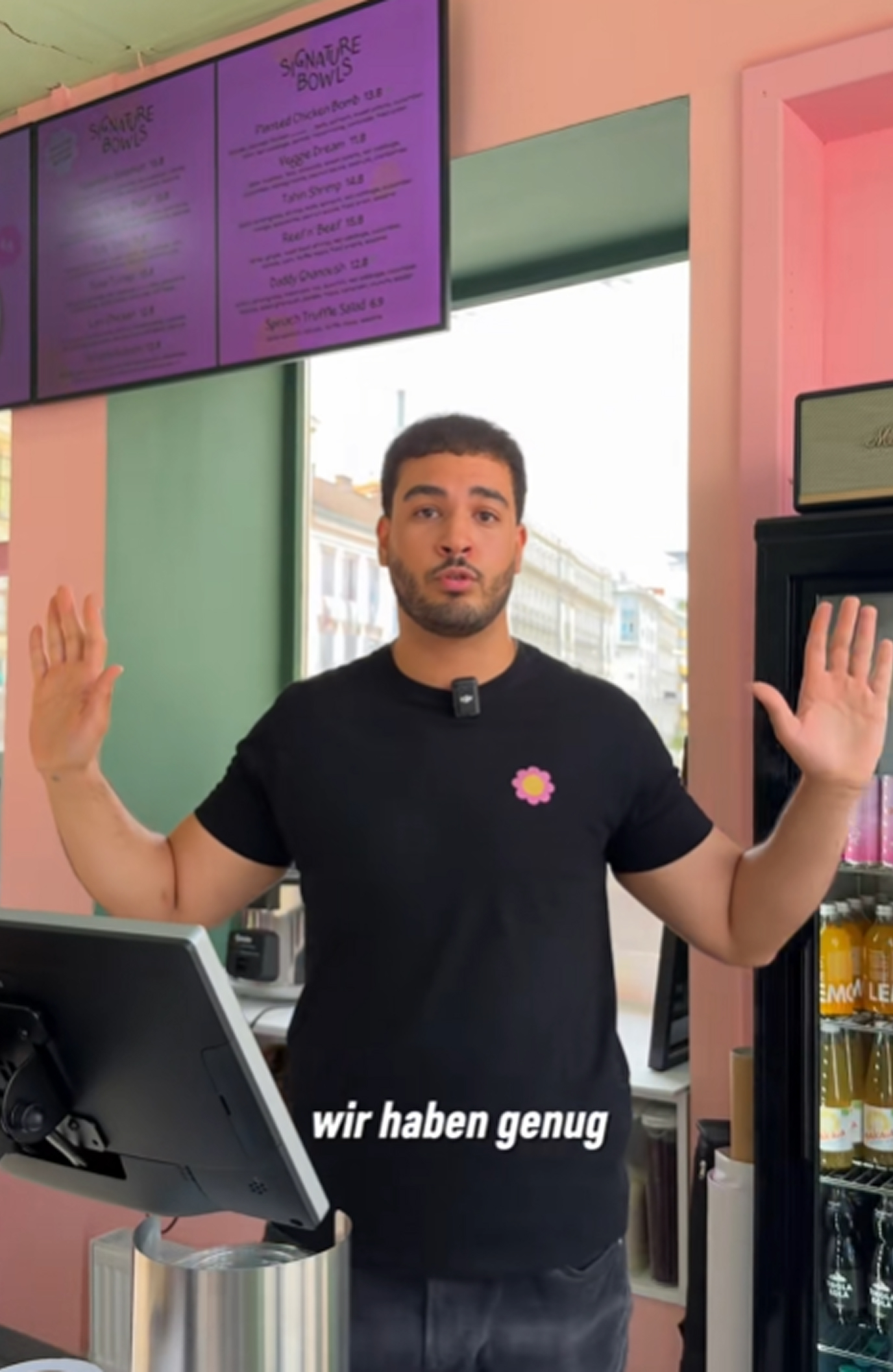 Man with raised hands standing behind a counter in a café or restaurant with a menu board above and a refrigerator with drinks to the side.
