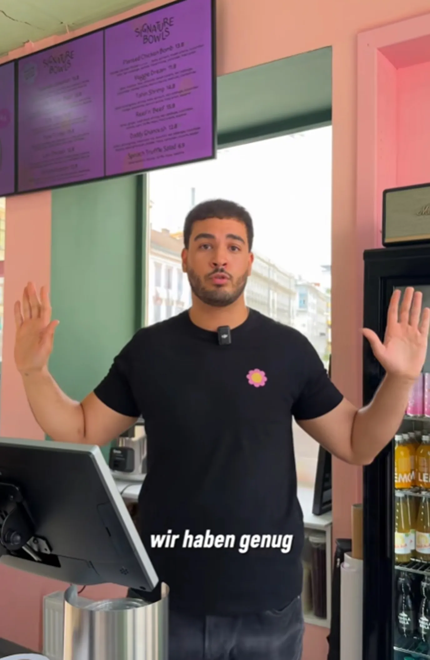 Man with raised hands standing behind a counter in a café or restaurant with a menu board above and a refrigerator with drinks to the side.