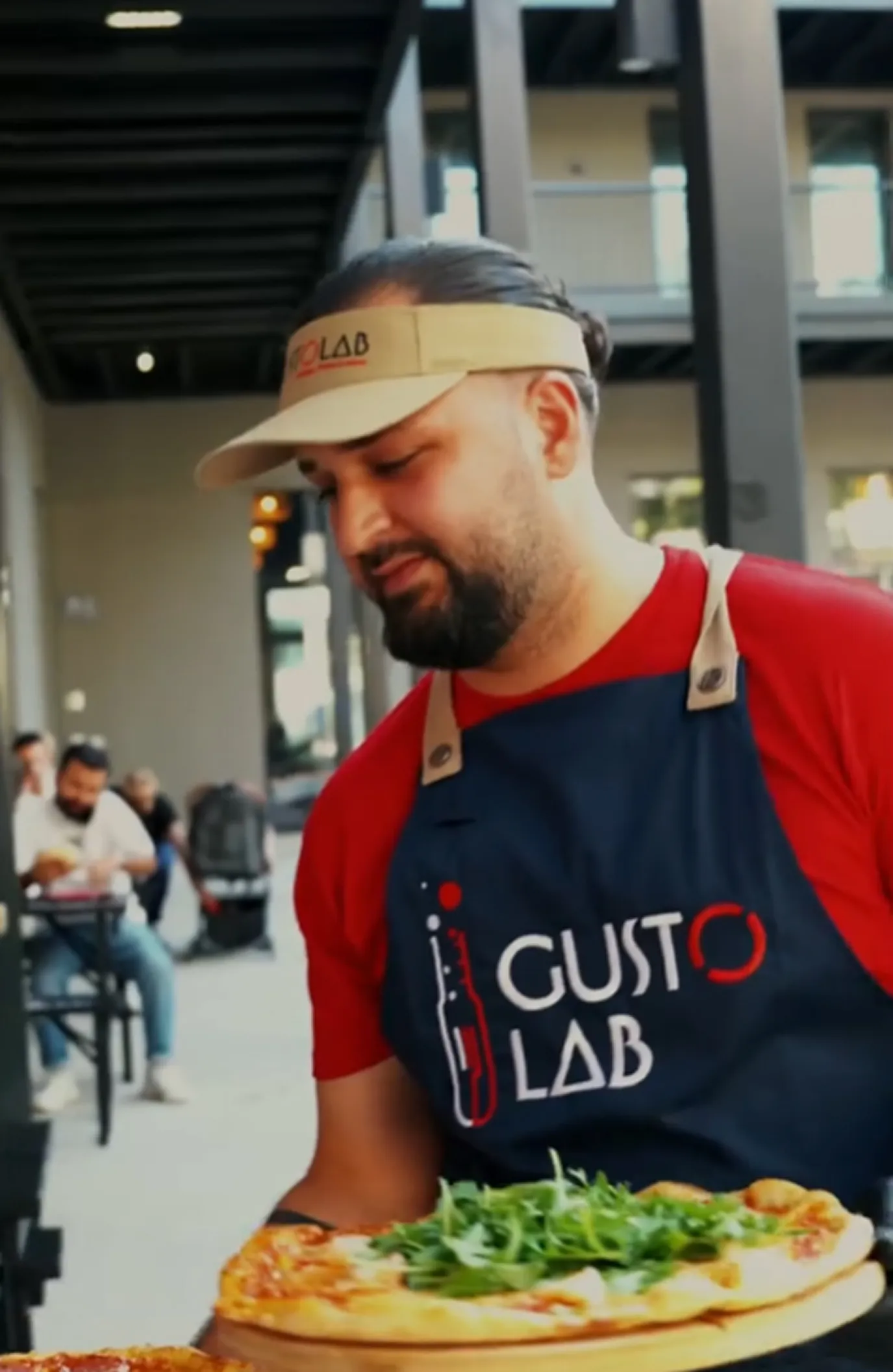 Man wearing a Gusto Lab apron and visor holding a pizza topped with arugula in an outdoor seating area.