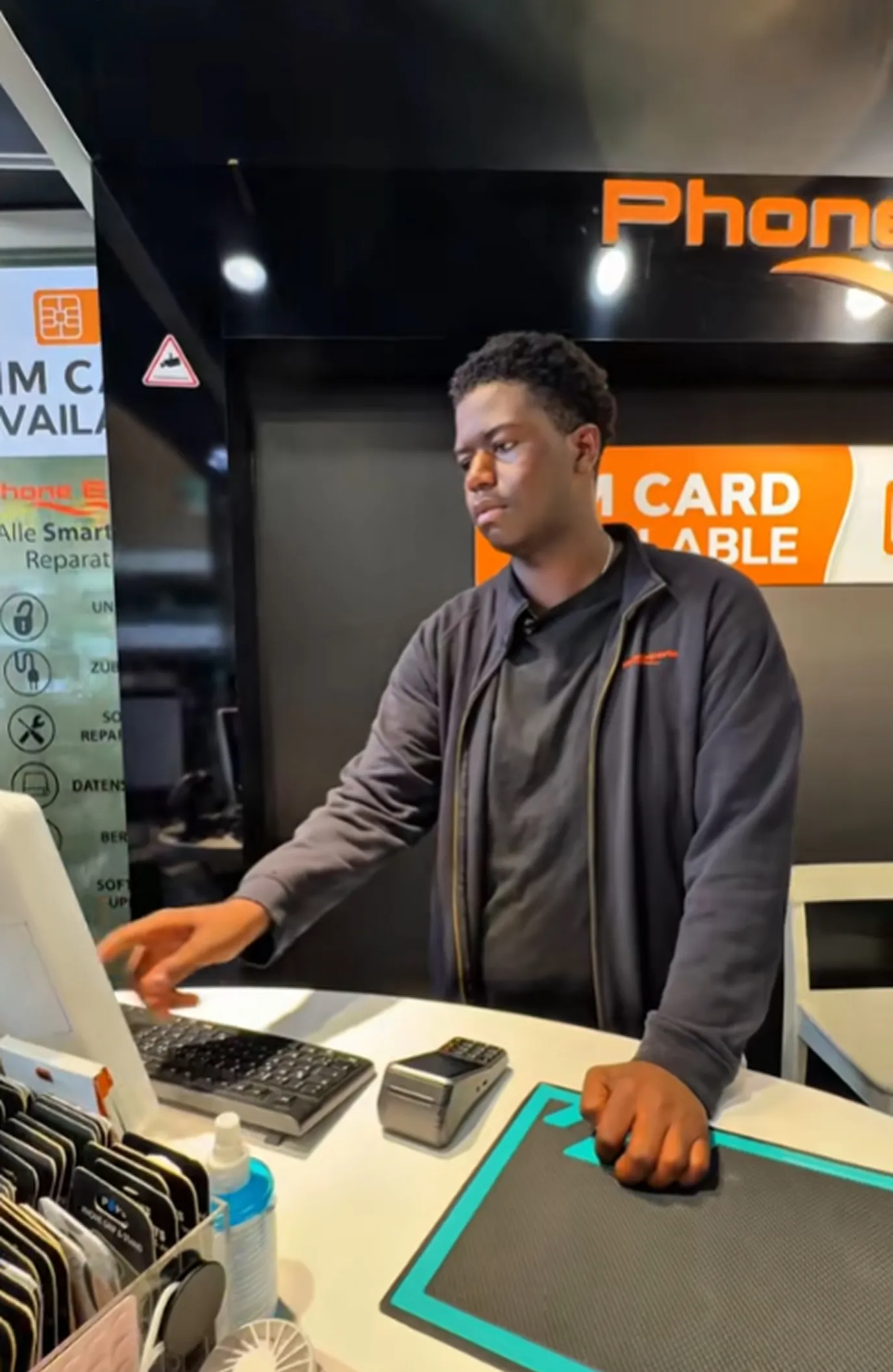 Young man working at a checkout counter with a keyboard, card reader, and display of phone accessories.