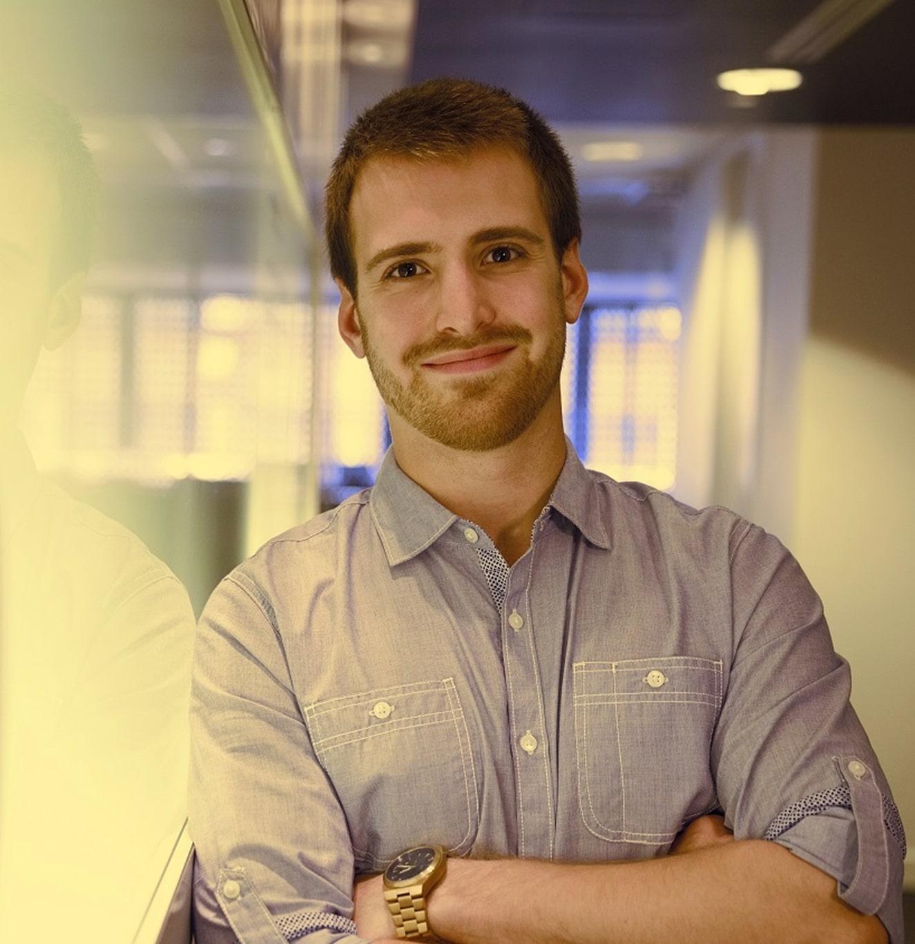 Smiling young man with short hair and beard standing with arms crossed near a glass wall indoors.