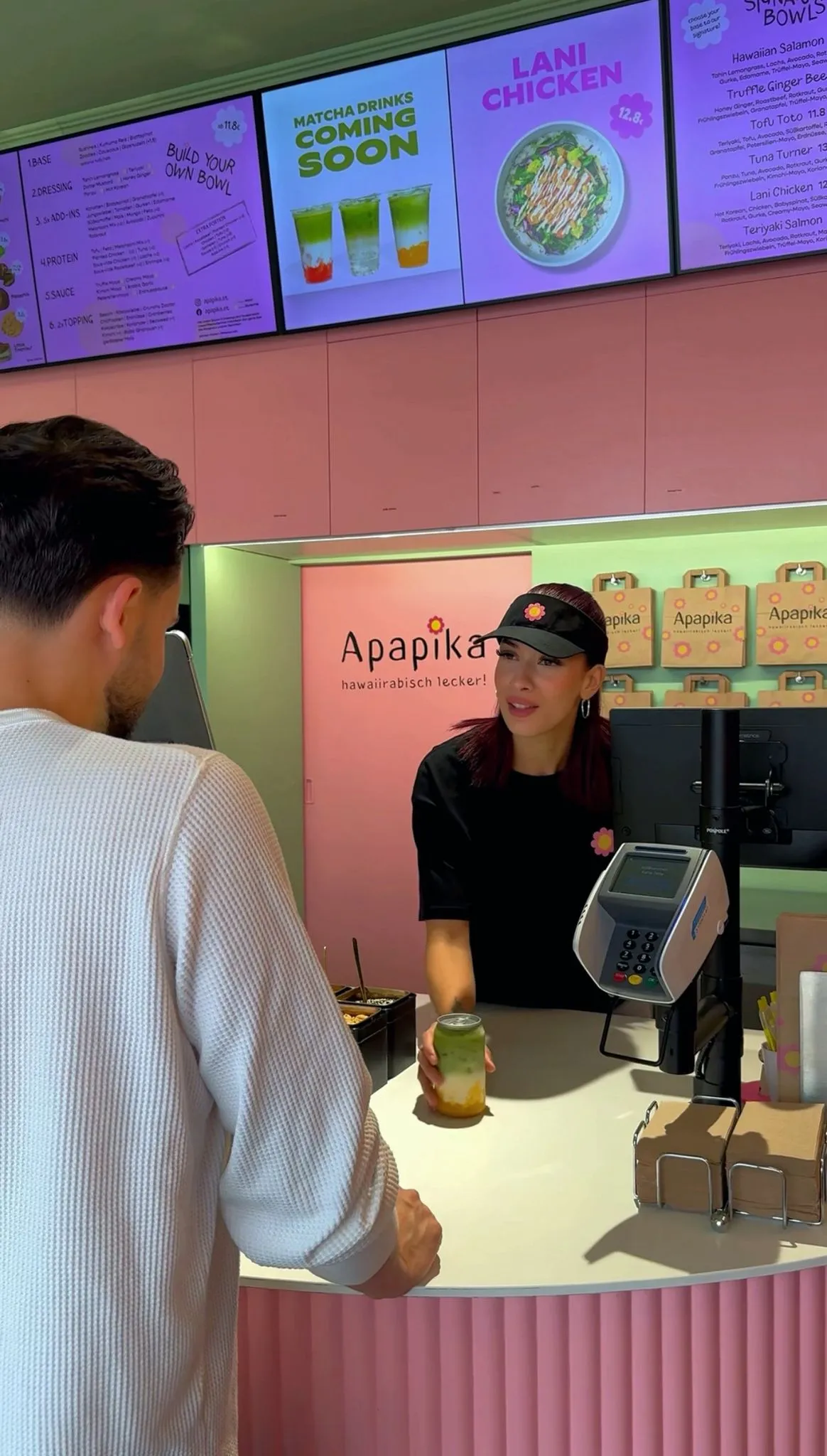 A woman barista handing a layered green and orange drink to a male customer at a pink-themed Apapika Hawaiian food and drink counter.