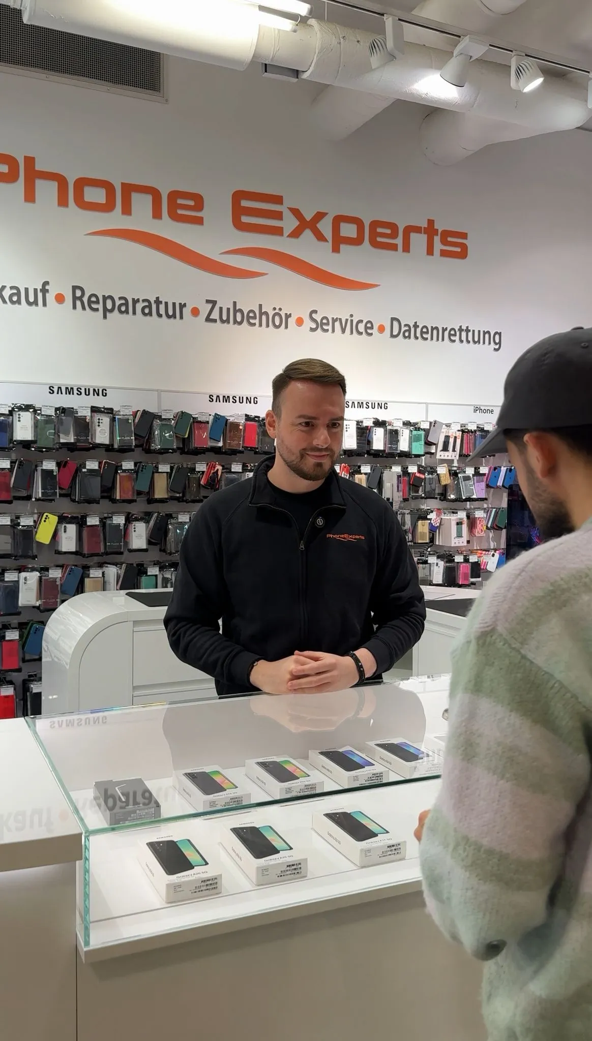A phone store employee in a black PhoneExperts jacket talks to a customer across a glass counter displaying Samsung smartphones in boxes.
