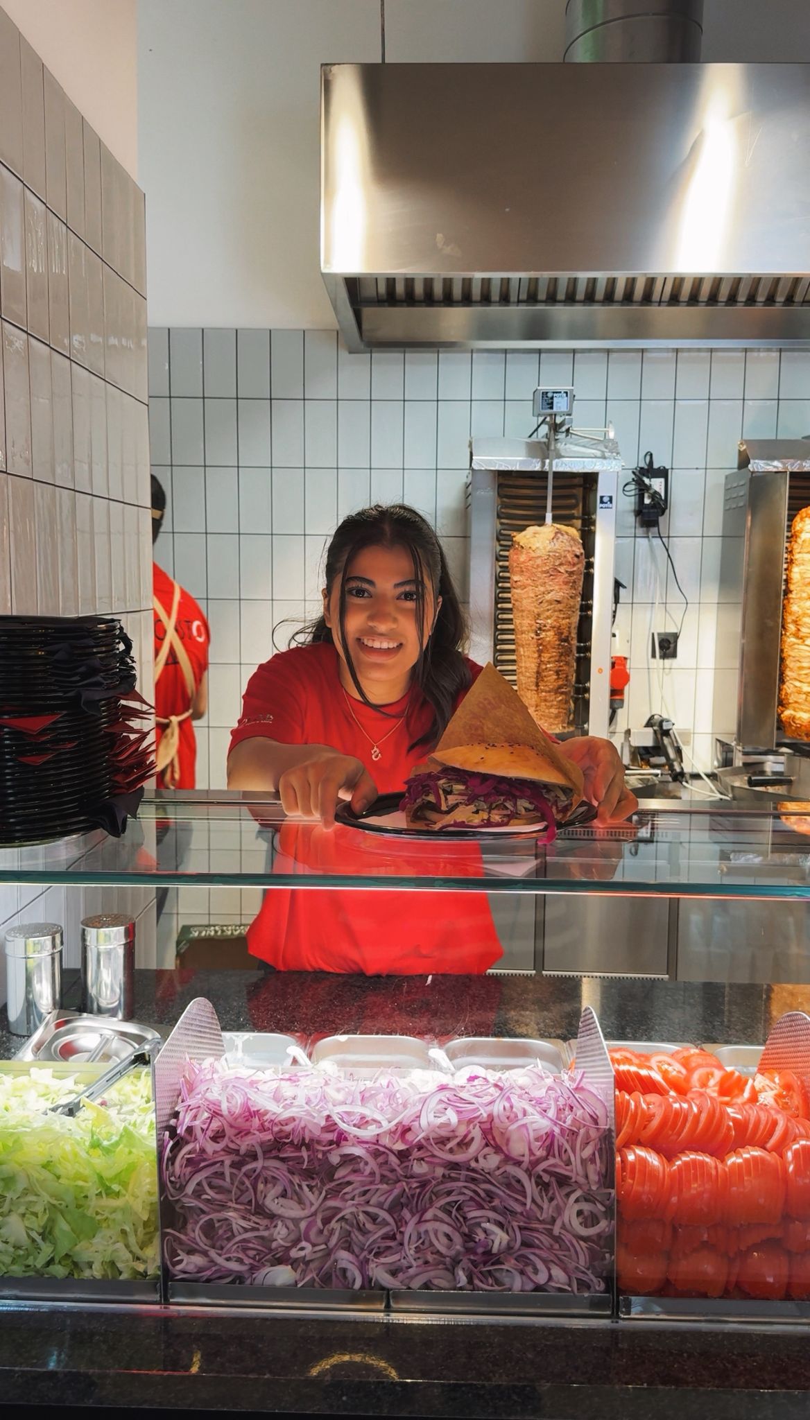 Smiling woman in a red shirt serving a plate with a wrap sandwich at a food counter with sliced onions, tomatoes, and lettuce in compartments.