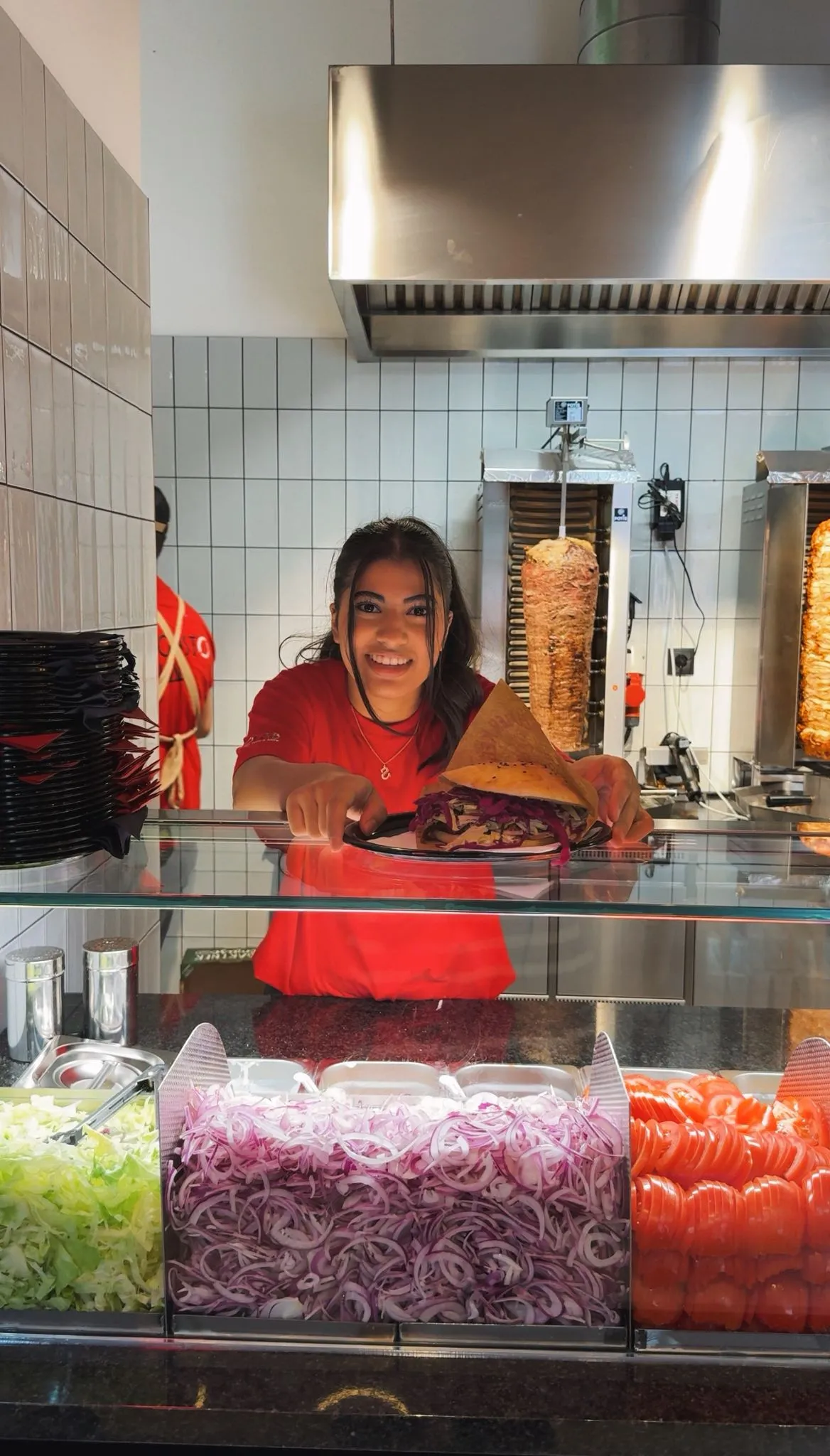 Smiling woman in a red shirt serving a plate with a wrap sandwich at a food counter with sliced onions, tomatoes, and lettuce in compartments.