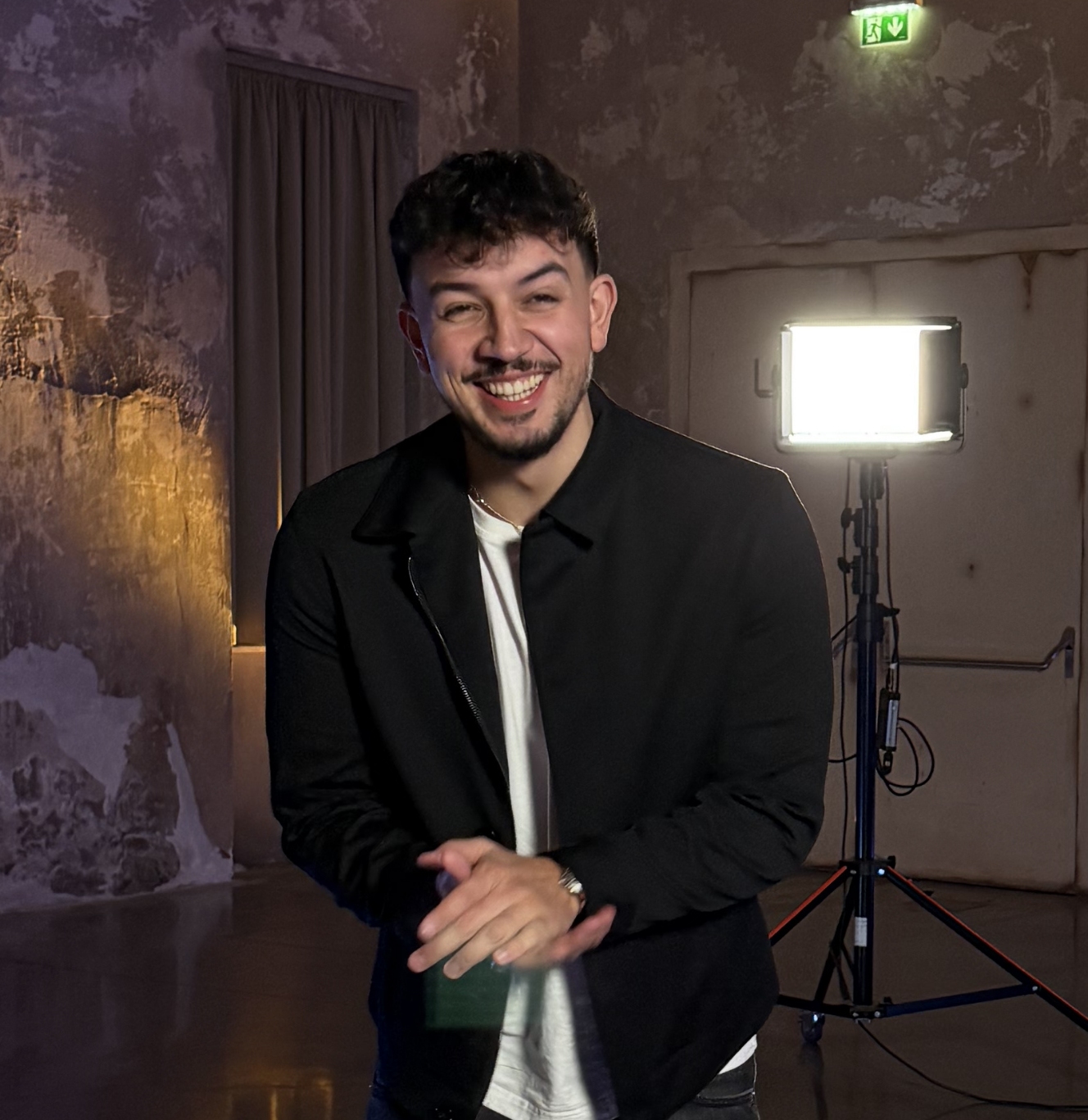 Smiling man with short curly hair and a beard wearing a black jacket and white shirt, standing indoors with bright studio light in the background.